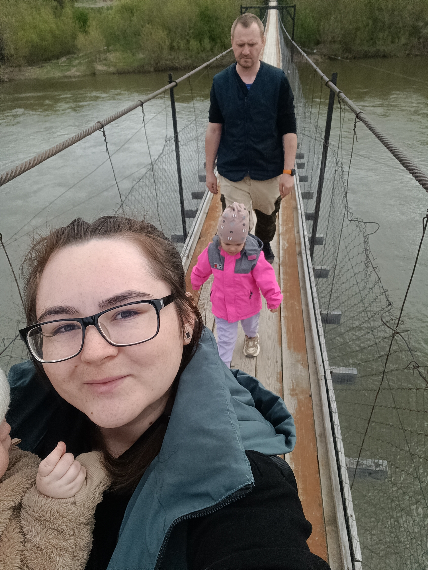 A family is walking on a suspension bridge over a river. The photograph is taken by a woman holding a baby, likely a selfie. Behind her is a toddler in a pink jacket and a man. The bridge is constructed of wooden planks, with metal cables acting as railings. The river below is murky. Trees are visible on the far bank. The location might be in or near the city of Посад, Russia.