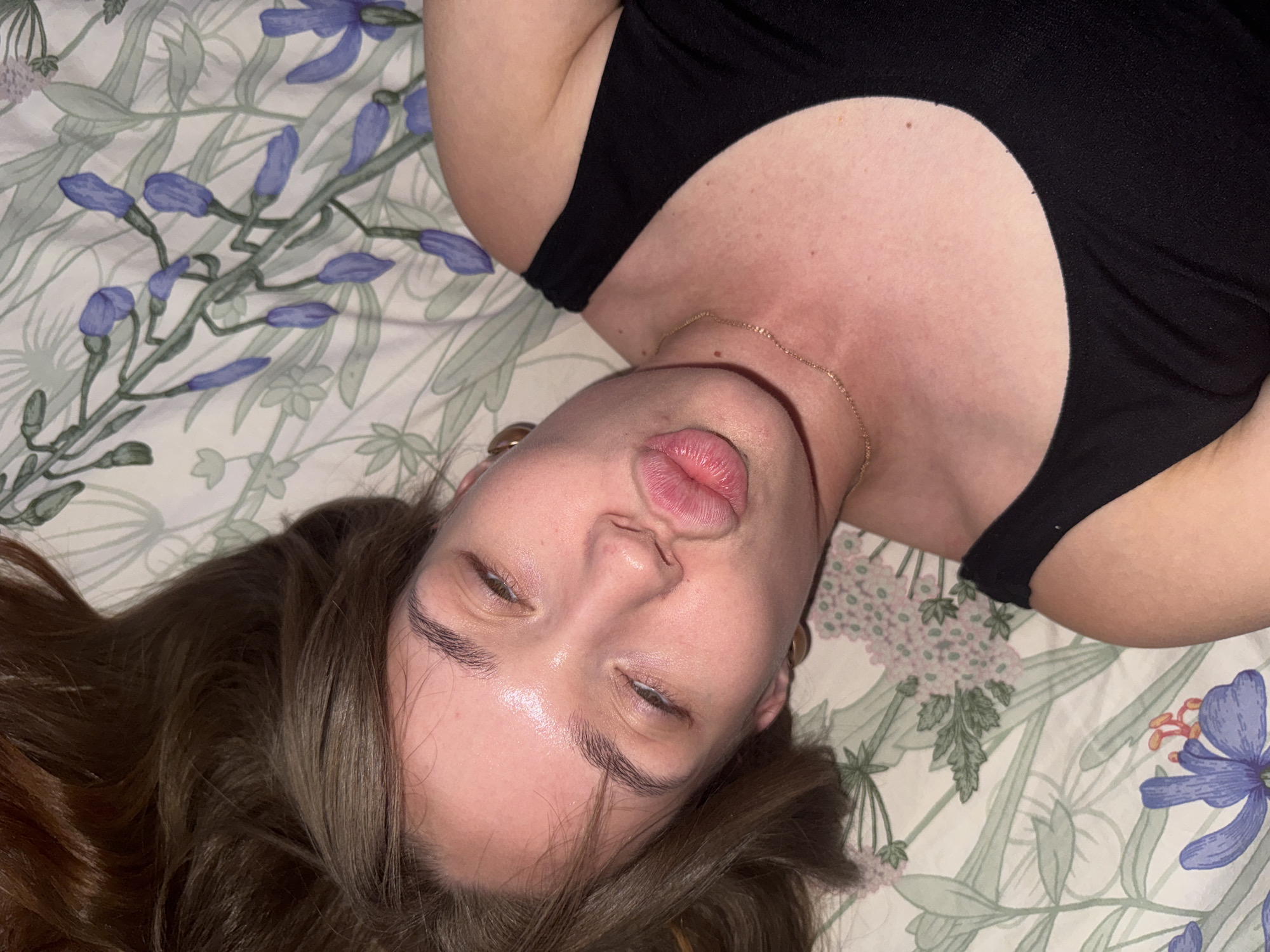 A young woman with long, brown hair is lying on her back on a bed with floral-patterned sheets. She is wearing a black tank top and a delicate gold necklace. Her eyes are closed, and her lips are pursed in a kiss. The lighting suggests an indoor setting, possibly a bedroom. There is no information in the image to suggest its location in Chaikovsky, Russia.