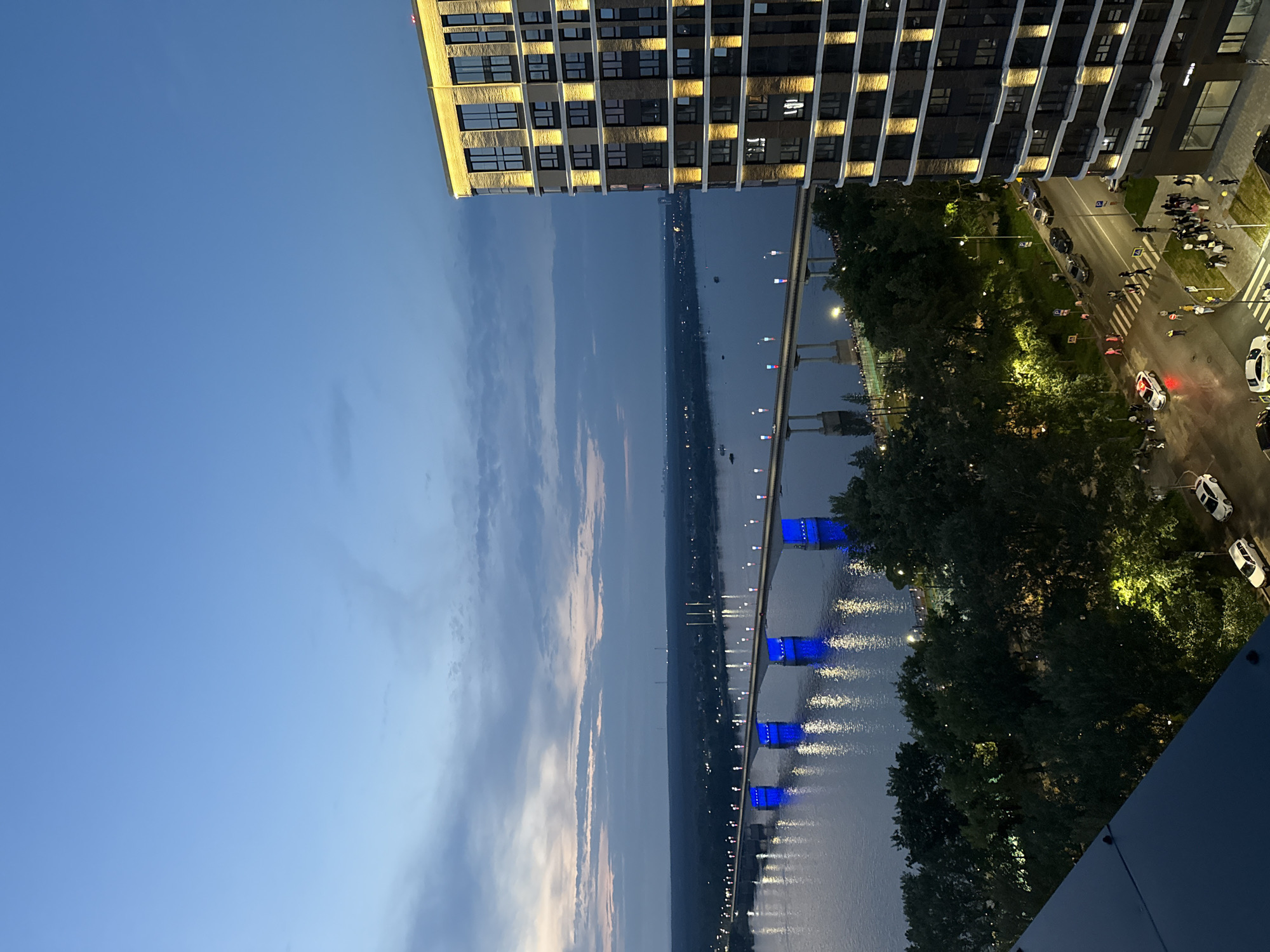 The image shows a panoramic view of the Kama River and a bridge in Perm, Russia, at dusk. A modern, multi-story building with illuminated windows stands prominently on the right side of the frame. The bridge is lit with blue lights, and its reflection is visible in the water. The sky is a deep blue with scattered clouds. Below the bridge, a road is visible with several cars and pedestrians. The scene appears to be a bustling urban environment.