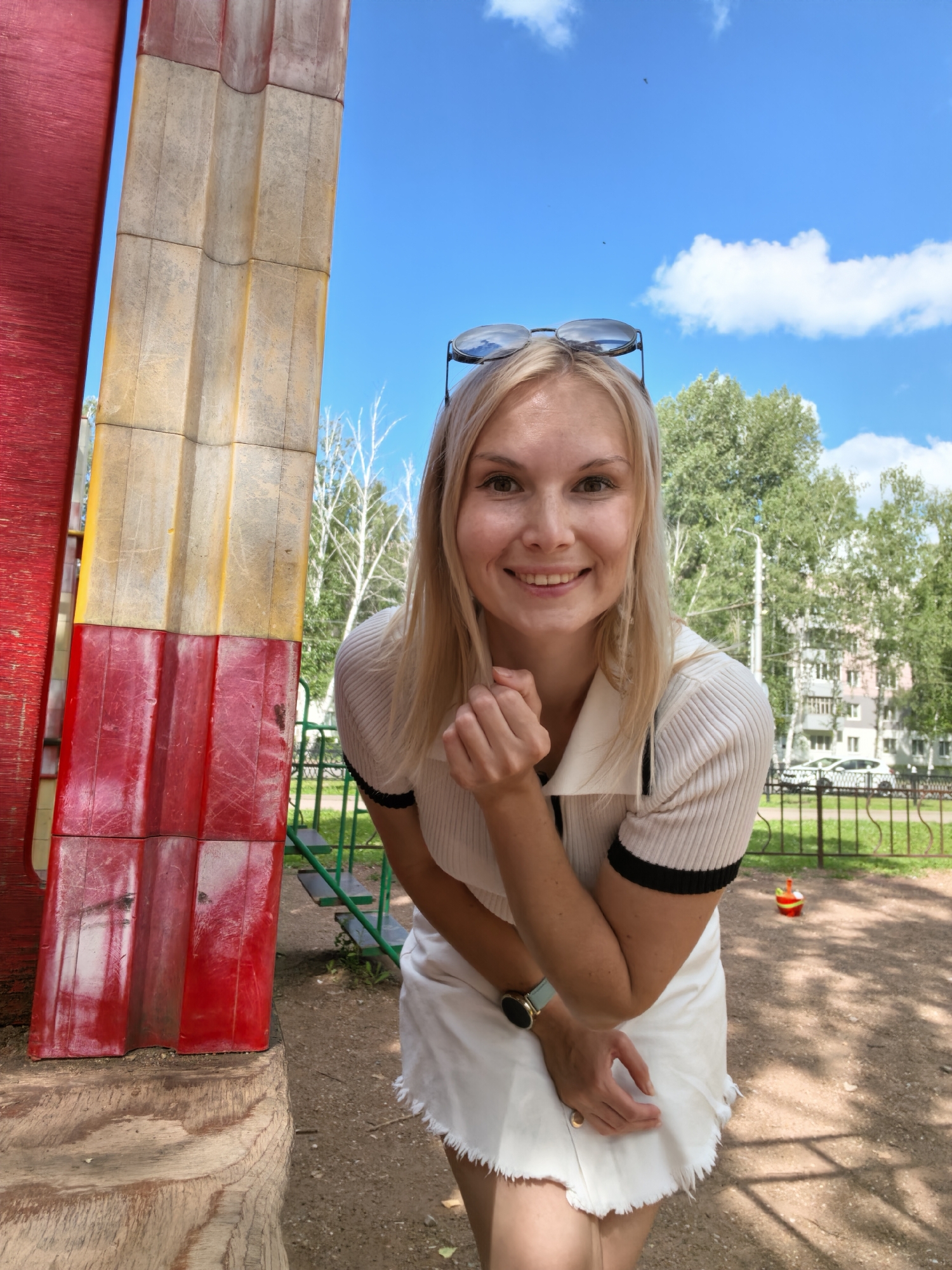 A blonde woman with fair skin and light makeup smiles at the camera. She is wearing a white short-sleeved shirt with a black collar and trim, and a white skirt. Sunglasses are perched on top of her head. She is standing in a playground with a large, colorful structure on her left. In the background, there are trees, a building, a fence, and playground equipment. The sky is blue with scattered white clouds. The ground is covered with dirt and sand. The woman's left hand is raised to her chin, and her right hand is resting on her skirt. The scene appears to be in an outdoor urban environment, possibly a residential area with a playground. It is not possible to determine the exact location within Sterlitamak, Russia from the image alone.