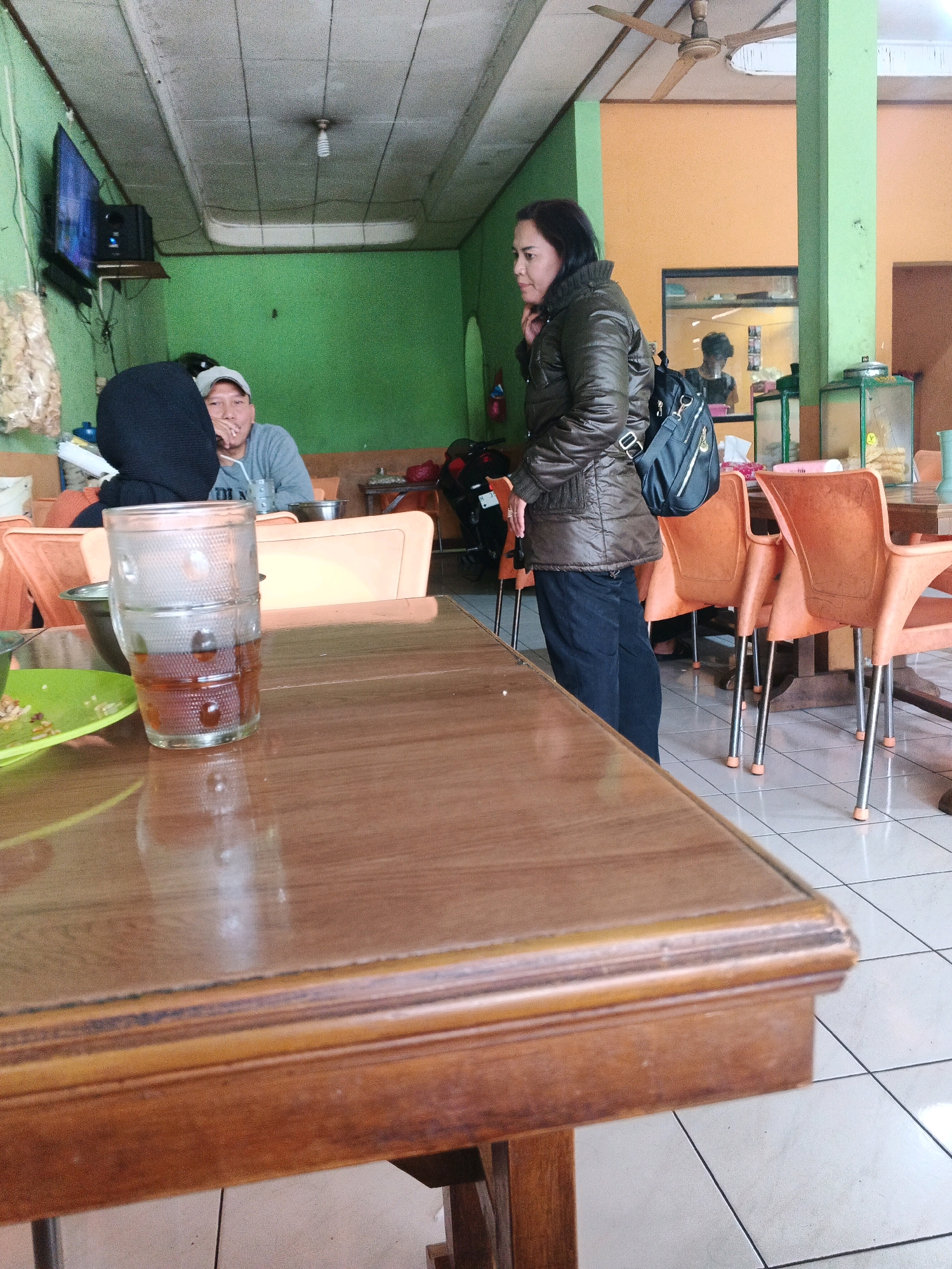 The image depicts an indoor scene, likely a casual eatery or restaurant in Ungaran, Indonesia. In the foreground, a wooden table with a polished surface is visible. On the table, a tall glass filled with a dark liquid, possibly tea or coffee, sits next to a small green plate with remnants of food.
To the left, a person wearing a black hijab is partially visible, seated at a table. Beside them, a man in a grey t-shirt and a baseball cap is also seated, holding a straw in his drink. In the middle ground, a woman stands facing away from the camera, wearing a dark, quilted jacket, dark pants, and a black backpack slung over her shoulder. She is positioned near a row of orange plastic chairs with metal legs, some of which are occupied by other patrons.
The environment features green walls and an orange wall in the background. A ceiling fan is mounted on the angled ceiling, and fluorescent lighting is visible. A television screen is mounted on the green wall to the left. Reflections in a mirror behind the standing woman suggest another room or area of the establishment. A motorcycle is parked to the far left, partially obscured. The floor is tiled in a white grid pattern. The overall impression is of a relaxed, everyday gathering place.