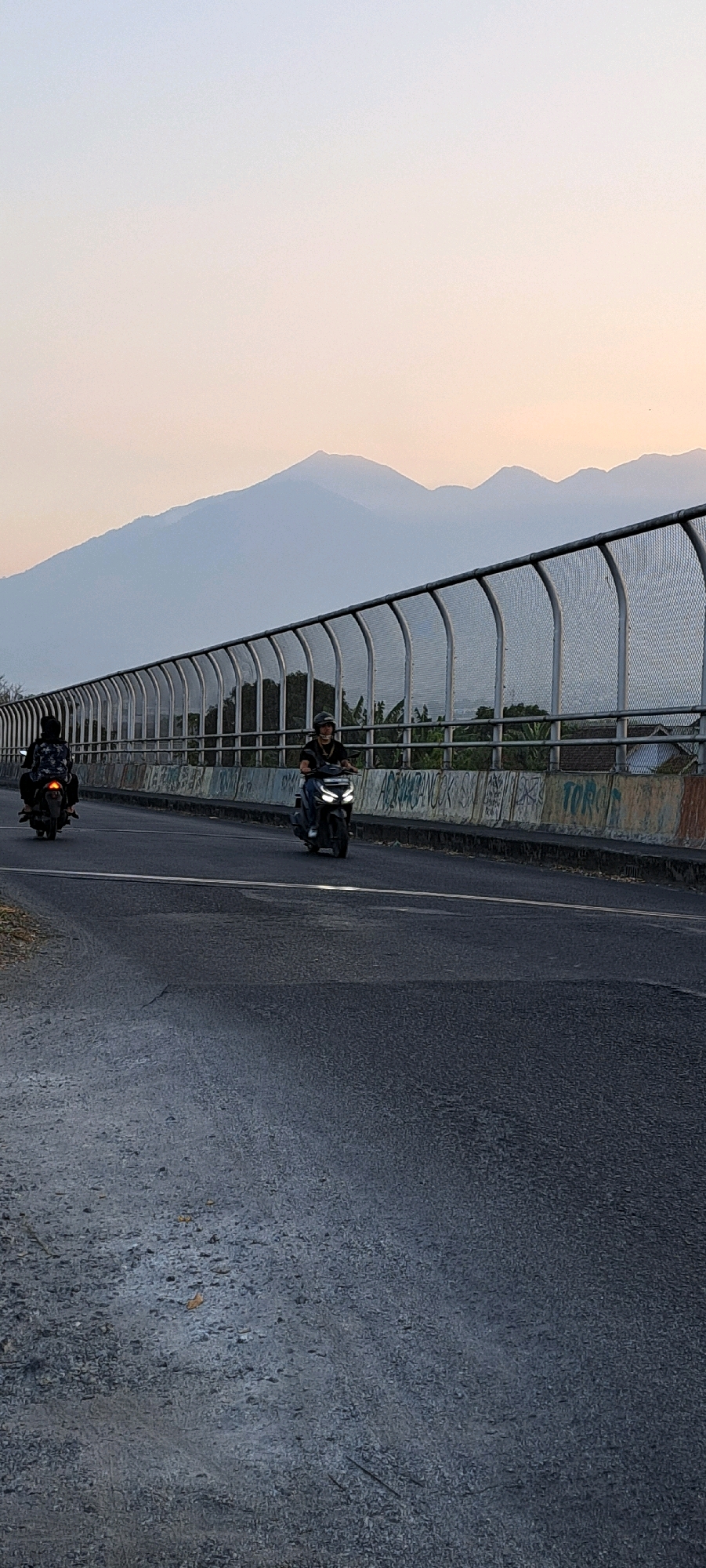 This is a vertical, eye-level, outdoor, daytime photograph taken from a road, possibly a bridge. The scene is dominated by a mountainous landscape in the background under a soft, hazy sky, transitioning from a pale orange at the horizon to a light blue. In the middle ground, a long, curved metal fence runs parallel to the road, creating a barrier. This fence has a mesh top and is supported by arched metal posts. Below the fence, a concrete barrier is visible, adorned with graffiti.
Two individuals on motorcycles are traveling on the road. The motorcycle on the left is further away and its rider is wearing a dark outfit. The motorcycle on the right is closer and its rider is wearing a dark shirt and a helmet, facing forward. Both motorcycles have their headlights on. The road surface appears to be asphalt, with some cracks and debris visible, particularly in the foreground. Trees and some structures are visible beyond the fence, suggesting a semi-urban or rural setting near a populated area. The overall atmosphere is serene, with the soft light indicating either early morning or late afternoon. The location is identified as Randupitu, Indonesia.