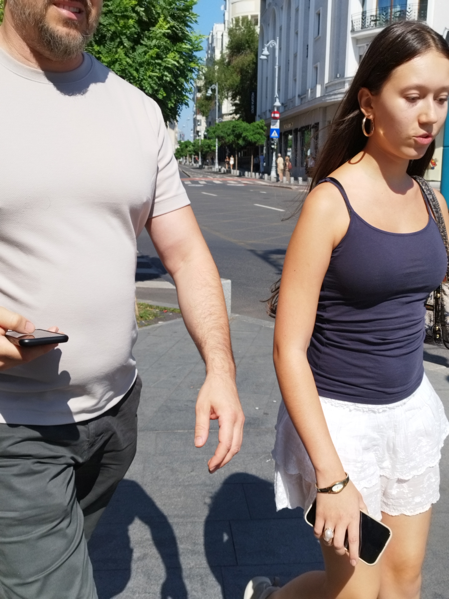 The image shows a man and a woman walking on a sidewalk in Bucharest, Romania. The man is on the left, wearing a light gray t-shirt and dark gray pants. He is holding a smartphone in his left hand. The woman is on the right, wearing a dark blue tank top and a white lace skirt. She is carrying a bag over her shoulder and holding a smartphone in her right hand. They are walking on a paved sidewalk next to a street with traffic. Buildings and trees line the street in the background. The lighting suggests it is daytime, likely sunny.
