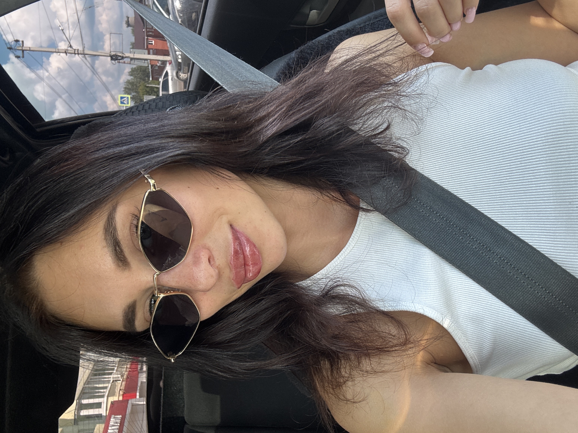 The image shows a young woman with dark hair and sunglasses, wearing a white tank top and a seatbelt, sitting in a car. The background outside the car window reveals a street scene in Chaikovsky, Russia. Buildings, power lines, traffic signals, trees, and other vehicles are visible, suggesting an urban or suburban environment. The woman is looking towards the camera, possibly taking a selfie.