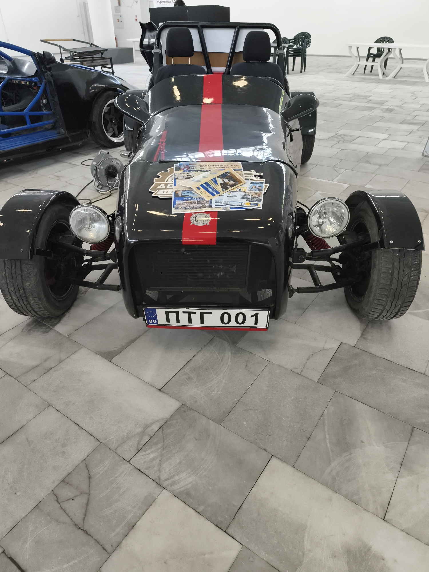 A black car with a red stripe and a license plate reading "PTG 001" is parked on a tiled floor. The license plate indicates that the car is from Bulgaria. There are several stickers on the front of the car. In the background, there are other vehicles and some tables and chairs. The text on the stickers indicates this image was captured in or pertains to the city of Targovishte, Bulgaria.