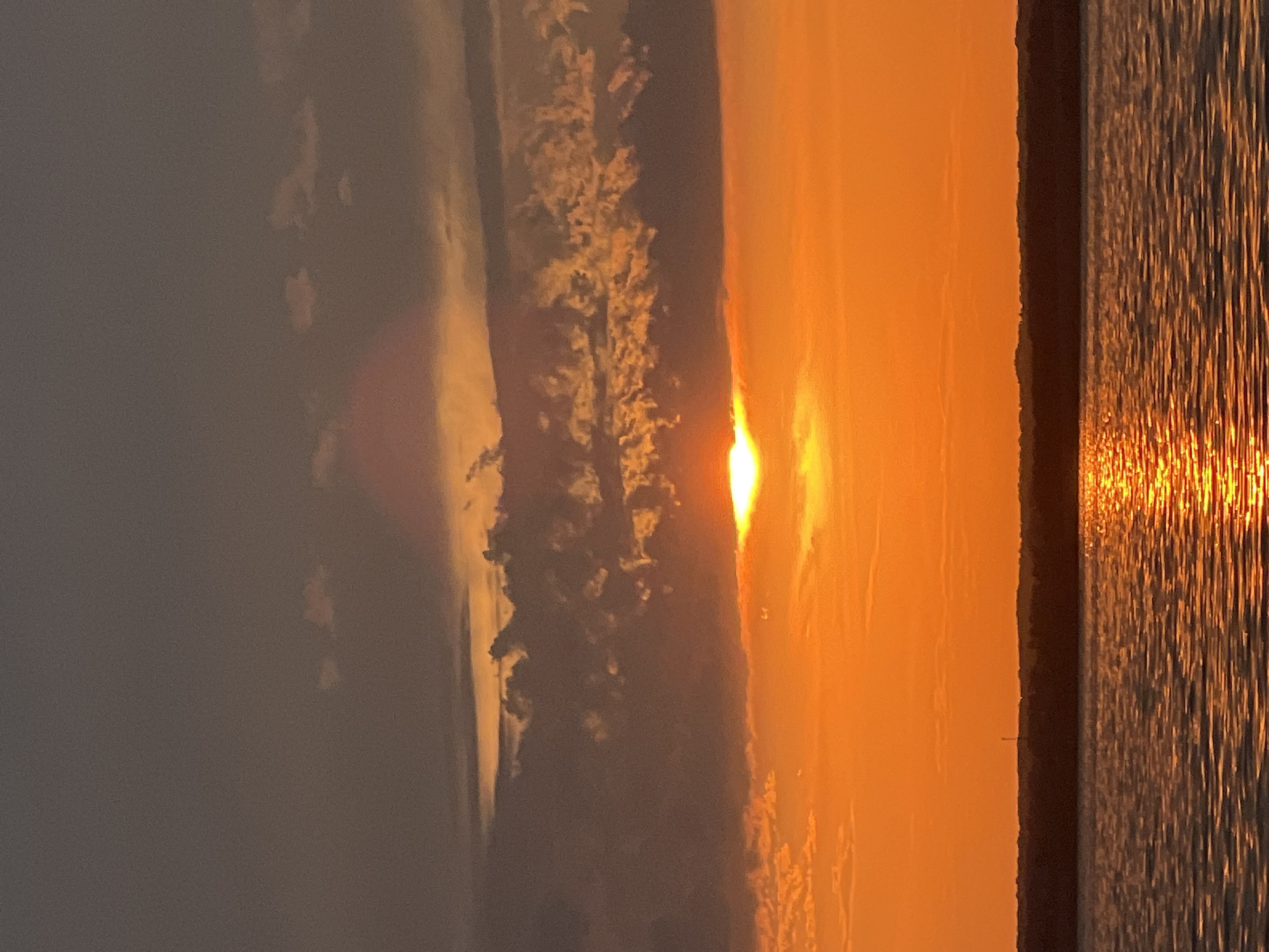 A tranquil sunset unfolds over the waters of Beregovoe, Russia. The sky is painted in hues of deep orange and gray, with fluffy clouds catching the sun's golden rays. The sun, a bright orb, is partially obscured by a band of clouds, casting a shimmering reflection on the calm water. A dark, silhouetted treeline stretches across the horizon, with a faint, thin tower visible in the distance. The foreground is dominated by the rippling surface of the water, reflecting the vibrant colors of the sky. There are no people or animals visible in this serene natural landscape.