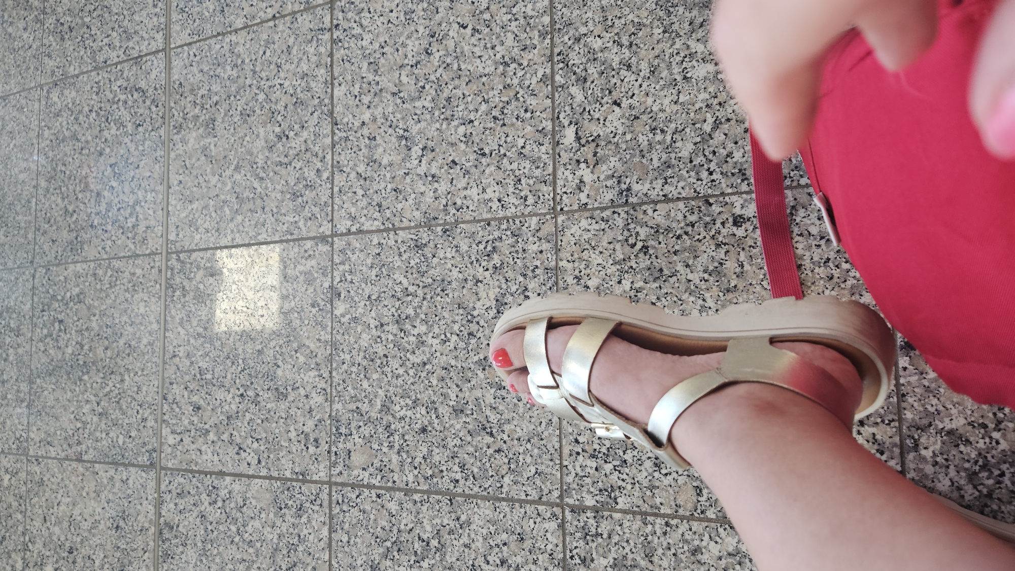 A close-up, high-angle shot shows a person's right foot wearing a gold sandal with a thick sole. The sandal has multiple straps across the foot, and the toes are painted with red nail polish. The foot is resting on a tiled floor with a speckled gray and black pattern. A bright red fabric, possibly clothing or a bag, is visible in the lower right corner of the frame, with a thin red strap extending across the floor. A blurred hand and fingers are in the foreground on the right. The lighting suggests an indoor environment, with a bright rectangular reflection on the floor tiles in the upper middle section of the image.