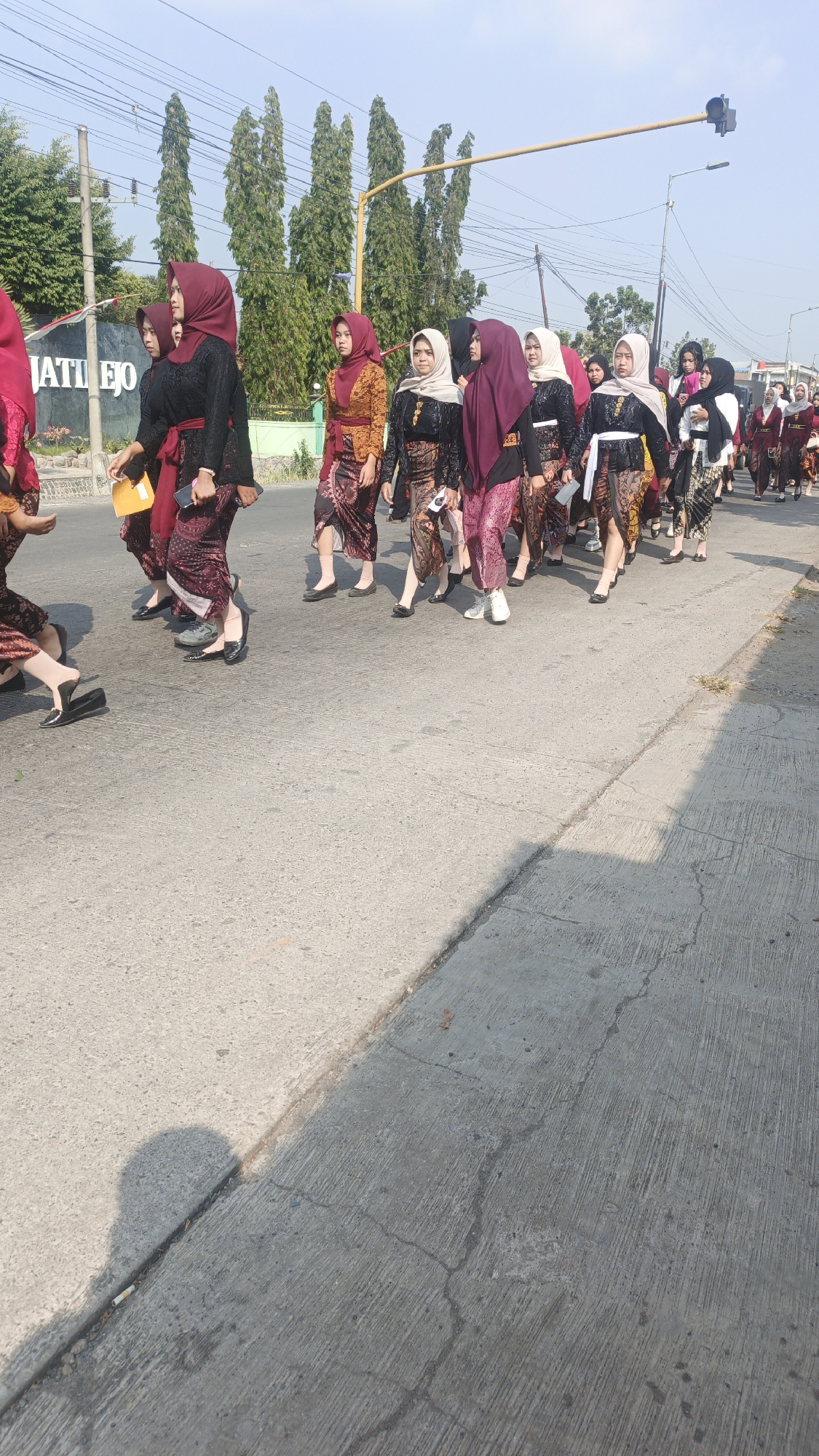 A group of young women, adorned in traditional Indonesian attire, are walking down a paved street under a clear blue sky. They are dressed in sarongs and black or brightly colored blouses, with headscarves in various hues. Some of the women are carrying yellow folders or cell phones. The street appears to be in a semi-urban or rural area, with trees lining the side of the road and a traffic light overhead. A sign that reads "JATI EJO" is visible on the left side of the image, suggesting the location is Jati Ejo, Sumengko, Indonesia. The ground is cracked and weathered, indicating it is an outdoor, public space. The overall impression is of a procession or cultural event.
