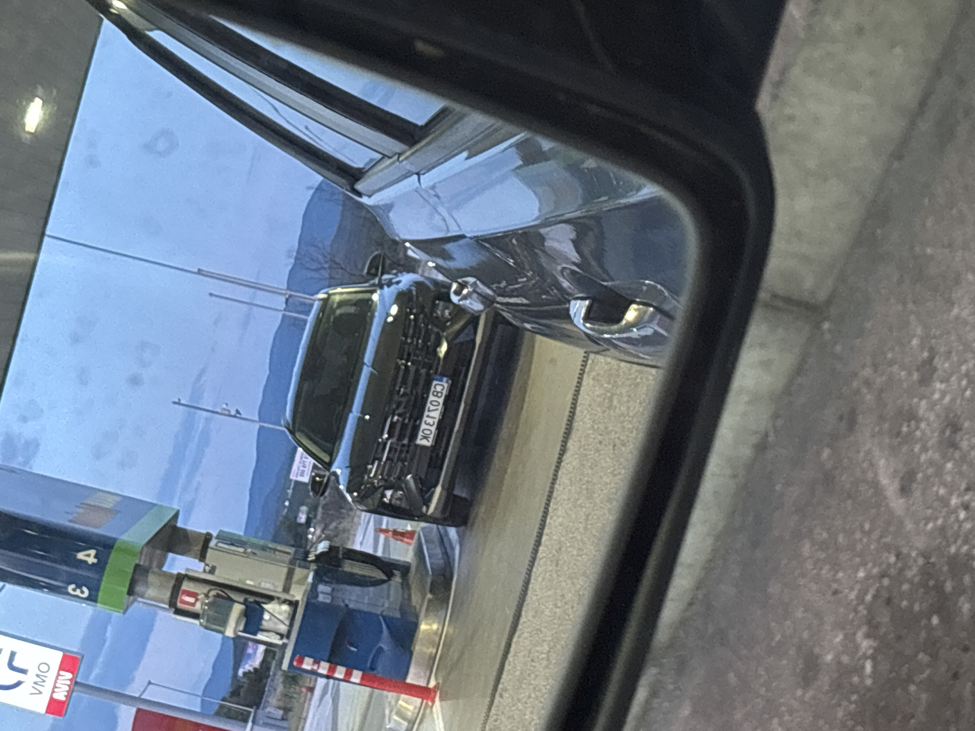 The image captures a scene at a gas station, likely in Kalugerovo, Bulgaria, as suggested by the context provided. The foreground is dominated by the reflection in a car's side mirror, showing the right side of the vehicle and the road. In the mid-ground, a black SUV is parked next to a blue gas pump. The SUV has its license plate visible, displaying the characters "KO E750 BO". The gas pump itself is tall and blue, with red and white striped posts nearby, and a red fire extinguisher mounted on its side. The background reveals a slightly hazy sky with scattered clouds and distant hills. The lighting suggests it is either dusk or early morning. There are no visible people in the frame.