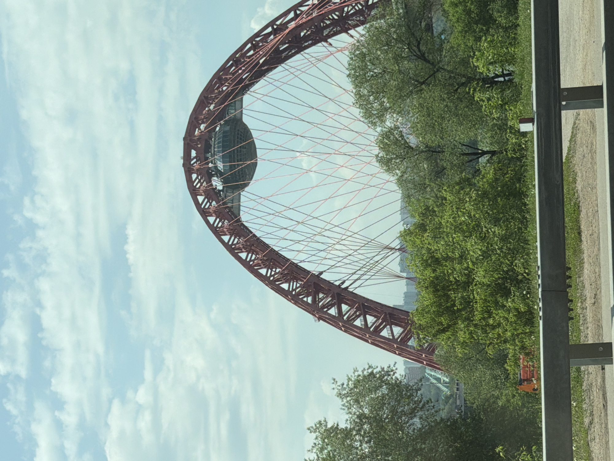 The image depicts a view of the Zhivopisny Bridge in Moscow, Russia. The bridge's distinctive red arch and spherical observation deck are prominently featured against a partly cloudy sky. Green trees and foliage surround the bridge structure. A metallic barrier or guardrail is visible in the foreground along with a grey gravel and grass embankment. An orange colored construction vehicle or equipment is visible through the foliage.