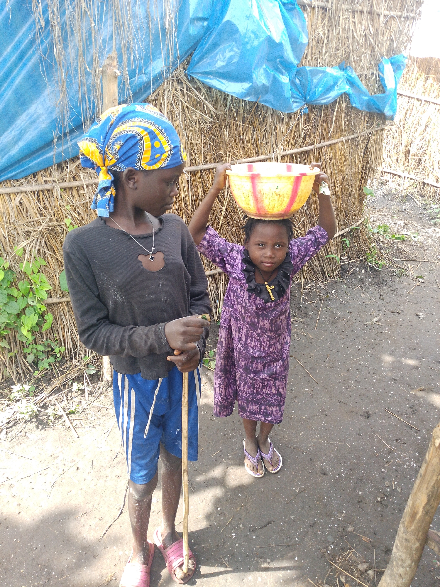 In Otulu, Nigeria, two young girls stand in front of a rustic dwelling constructed from thatch and covered with a blue tarp. The older girl, wearing a blue and yellow headscarf, a black long-sleeved shirt, and blue shorts with white stripes, holds a wooden stick. She looks to her right, her expression pensive. The younger girl, dressed in a purple patterned dress, balances a yellow and red bowl on her head. She wears a golden cross necklace. Her gaze is directed forward, her expression serious. The ground around them is dirt and debris.