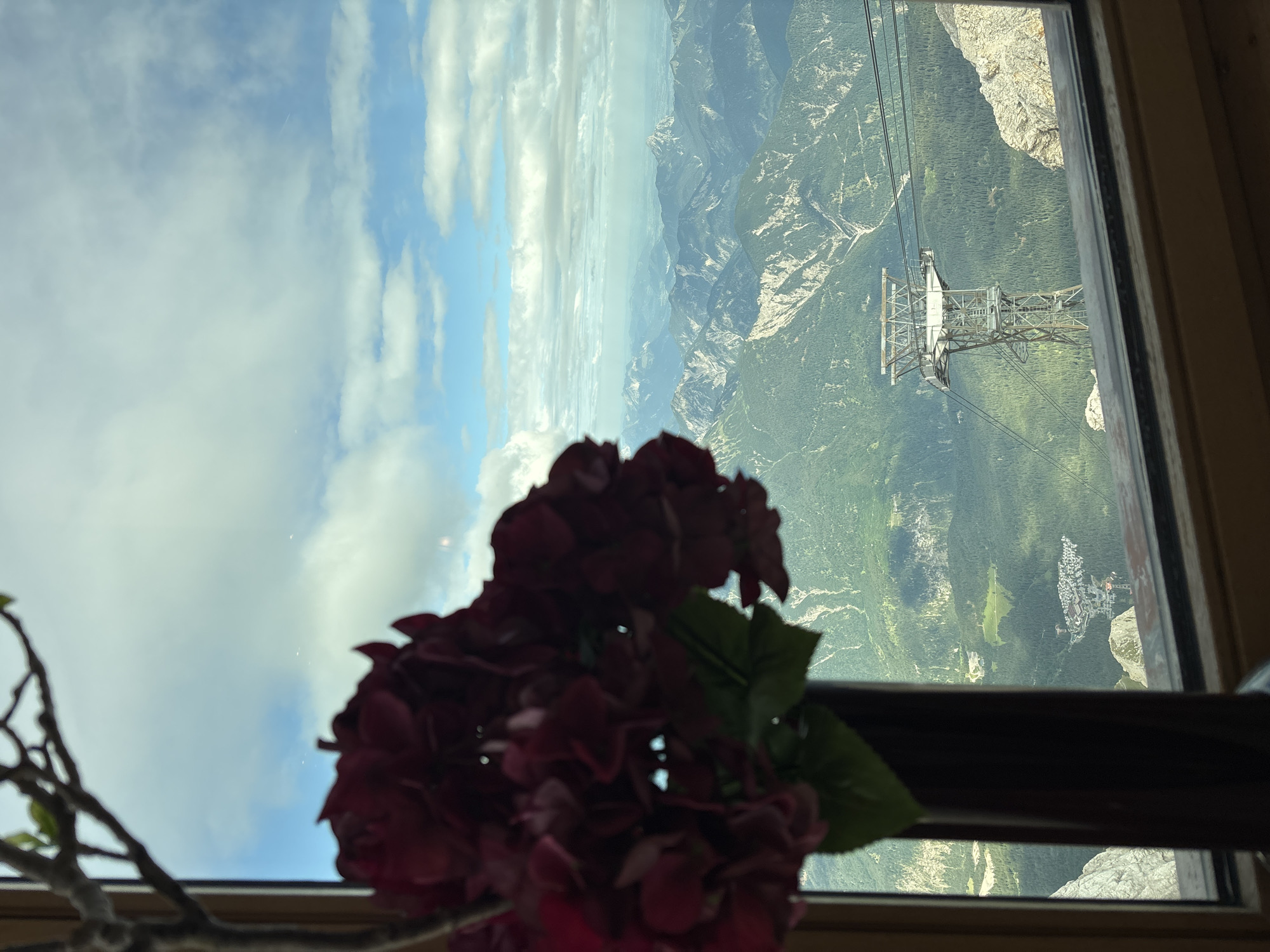 A view from a window in Ehrwald, Austria, looks out over a vast mountain landscape under a partly cloudy sky. In the foreground, a dark vase holds a cluster of deep red, artificial flowers, partially obscuring the lower left corner of the scene. The window frame, made of light brown wood, is visible along the bottom and left edge.
Beyond the flowers and window, layers of green, forested mountains recede into the distance, interspersed with rocky peaks. A prominent cable car tower, a steel structure with multiple cables stretching across the frame, stands in the mid-ground. Below and to the left of the tower, a small village or cluster of buildings is nestled amongst the trees. The sky is a mixture of bright blue and scattered white clouds, with some wispy formations stretching towards the horizon. The overall impression is one of serene natural beauty, framed by the interior of a building.