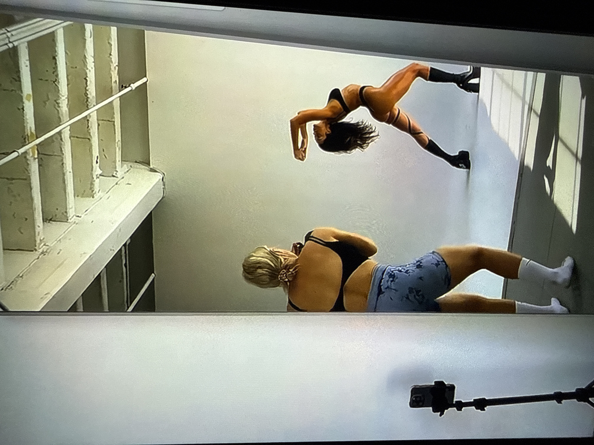 The image, likely a photograph of a digital display or a reflection, depicts an indoor photography studio located in Unknown, Unknown. The scene is illuminated by strong directional lighting, casting prominent rectangular shadows onto a white seamless backdrop that covers both the wall and floor. Above the backdrop, an industrial-style ceiling with exposed beams, what appear to be concrete blocks, and a white pipe running horizontally is visible.
In the foreground to the left, a person with blonde hair styled in a bun with a patterned scrunchie is seen from behind. They are wearing a black multi-strap sports bra, blue patterned athletic shorts, and white socks. They are in a slightly bent-knee stance, holding a camera (partially obscured) and actively photographing the model.
To the right, a model with dark, wavy hair is captured in a dynamic pose, arching their back significantly with arms bent and hands near their head, looking upwards and to their left. The model is wearing a black bikini-style top and bottom, and black high-heeled platform boots with decorative straps wrapped around their upper thighs. Their exposed skin appears glossy.
On the far left, a black tripod supports a dark-colored smartphone, oriented vertically and pointed towards the studio setup, presumably recording or capturing additional content.