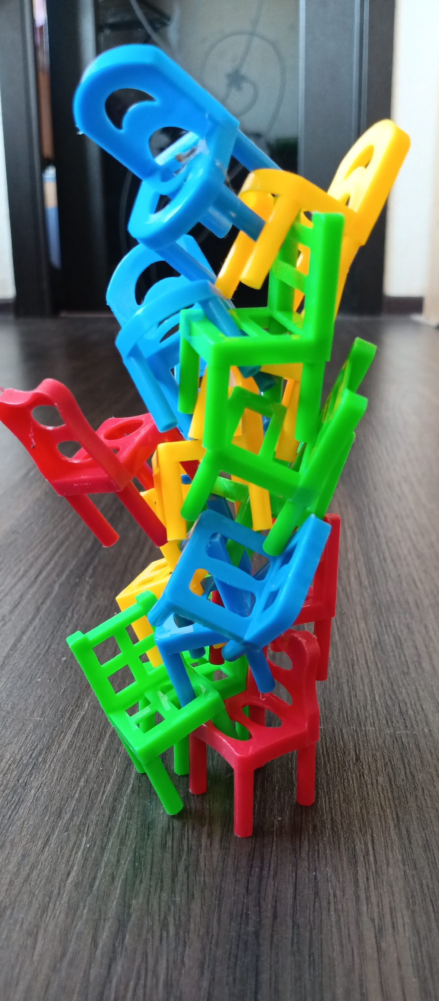A stack of colorful miniature plastic chairs, interlocked and precariously balanced, rests on a wood-grain surface. The chairs are primarily red, blue, green, and yellow. The setting appears to be indoors, with a blurred background showing a dark door and a light wall. The location is Unknown, Unknown.
