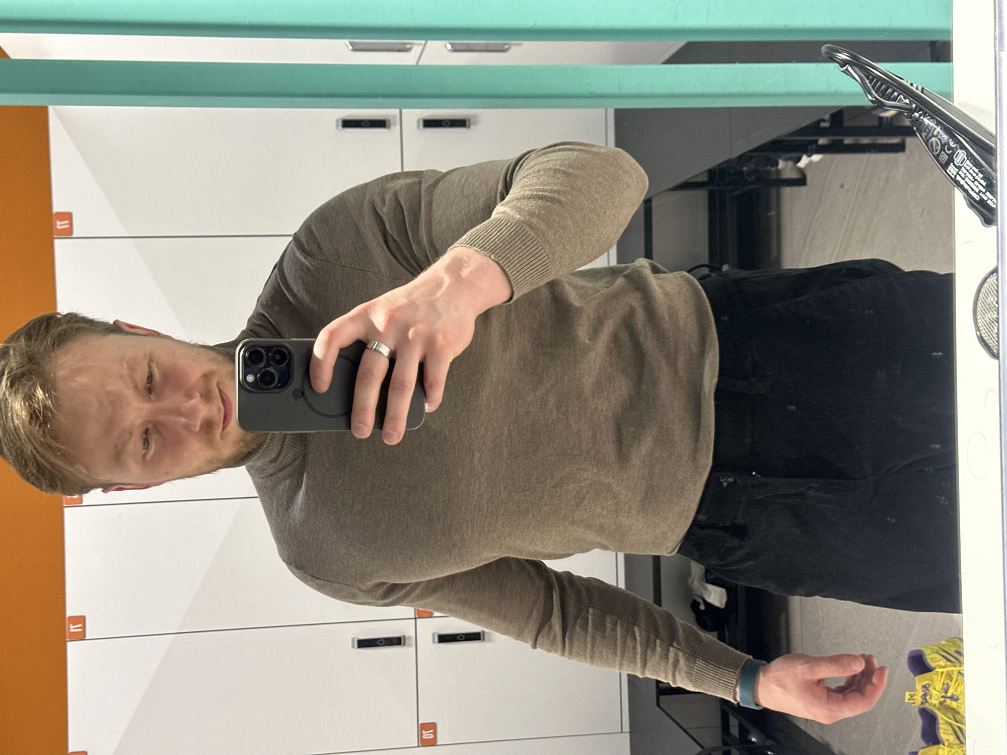 A man stands in front of a mirror in what appears to be a locker room. The lockers are white with orange ends and have numbers on them. The man is wearing a brown sweater and black pants. He is holding a cell phone in his right hand and taking a selfie. He is fair-skinned with blond hair. In the background, there is a pair of yellow shoes on the floor. The mirror is framed by a turquoise and white trim. There is no information to confirm that this image was taken in Sochi, Russia.