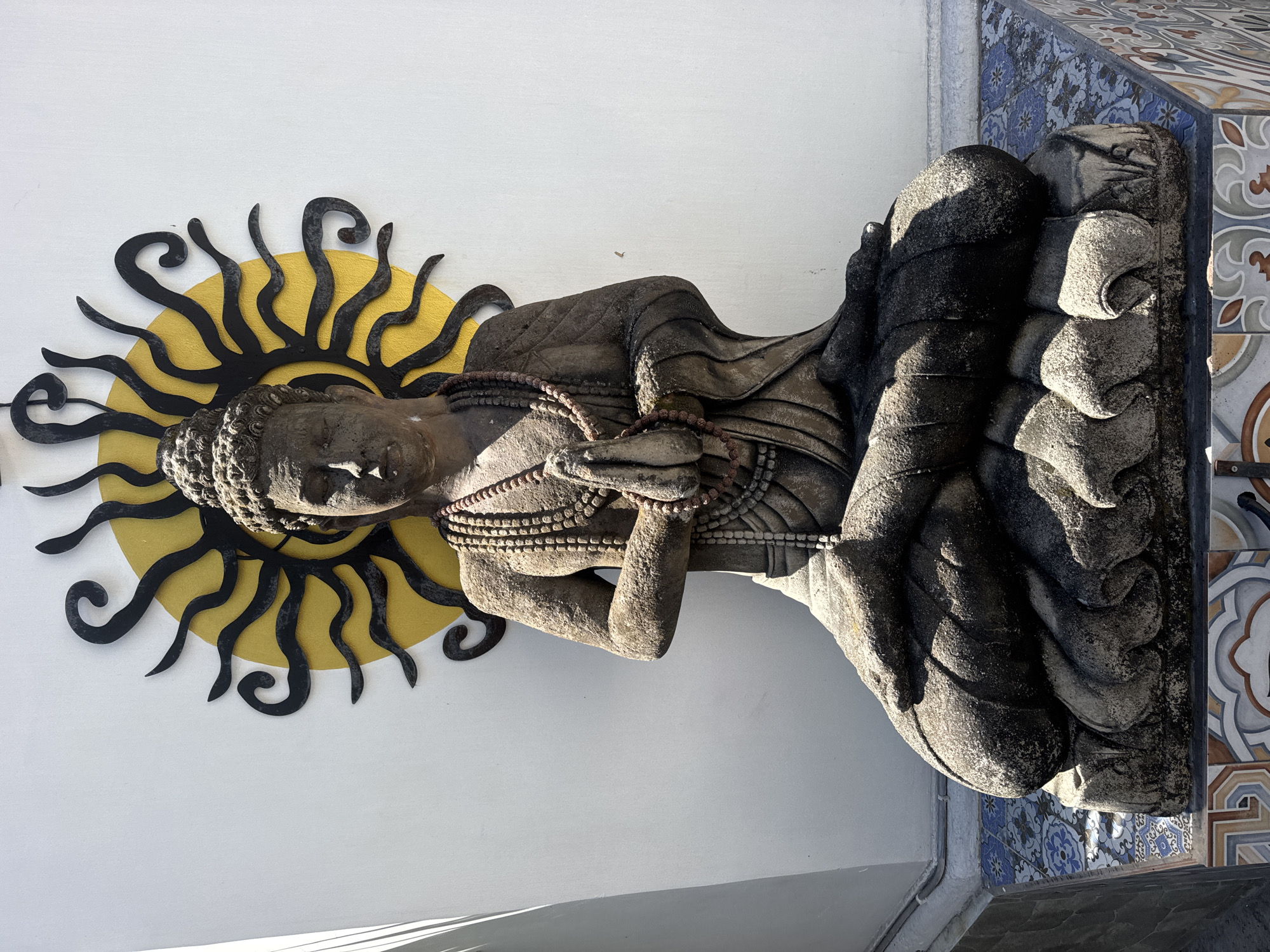 A stone statue of Buddha is depicted in a seated meditation pose, with hands pressed together in front of the chest, against a white wall. Behind the Buddha statue, a decorative sunburst motif with yellow and black elements is mounted on the wall. The statue rests on a base adorned with colorful, patterned tiles. The scene appears to be outdoors, with sunlight casting shadows. This statue is located in Kuta, Indonesia, at the entrance of the Dewa Ruci statue.