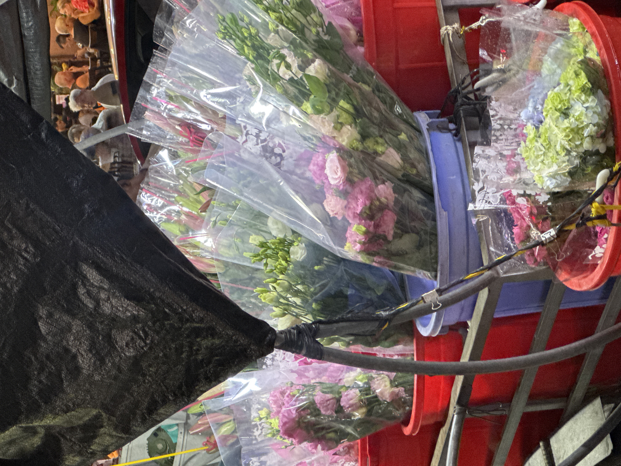 The image depicts a street-side flower stall in Bac Nha Trang Ward, Vietnam. The foreground is dominated by an array of flower bouquets, each individually wrapped in clear plastic. These bouquets vary in color, with prominent arrangements of pink roses and white and green lisianthus. The flowers are held in large plastic buckets, some red and some blue. A black tarp hangs from above, casting a shadow and obscuring the upper portion of the scene. In the background, a group of people are visible sitting at tables, suggesting a nearby establishment such as a restaurant or cafe. The lighting is dim, indicating it might be evening or nighttime. The overall impression is that of a bustling urban environment with a focus on the vibrant floral offerings.