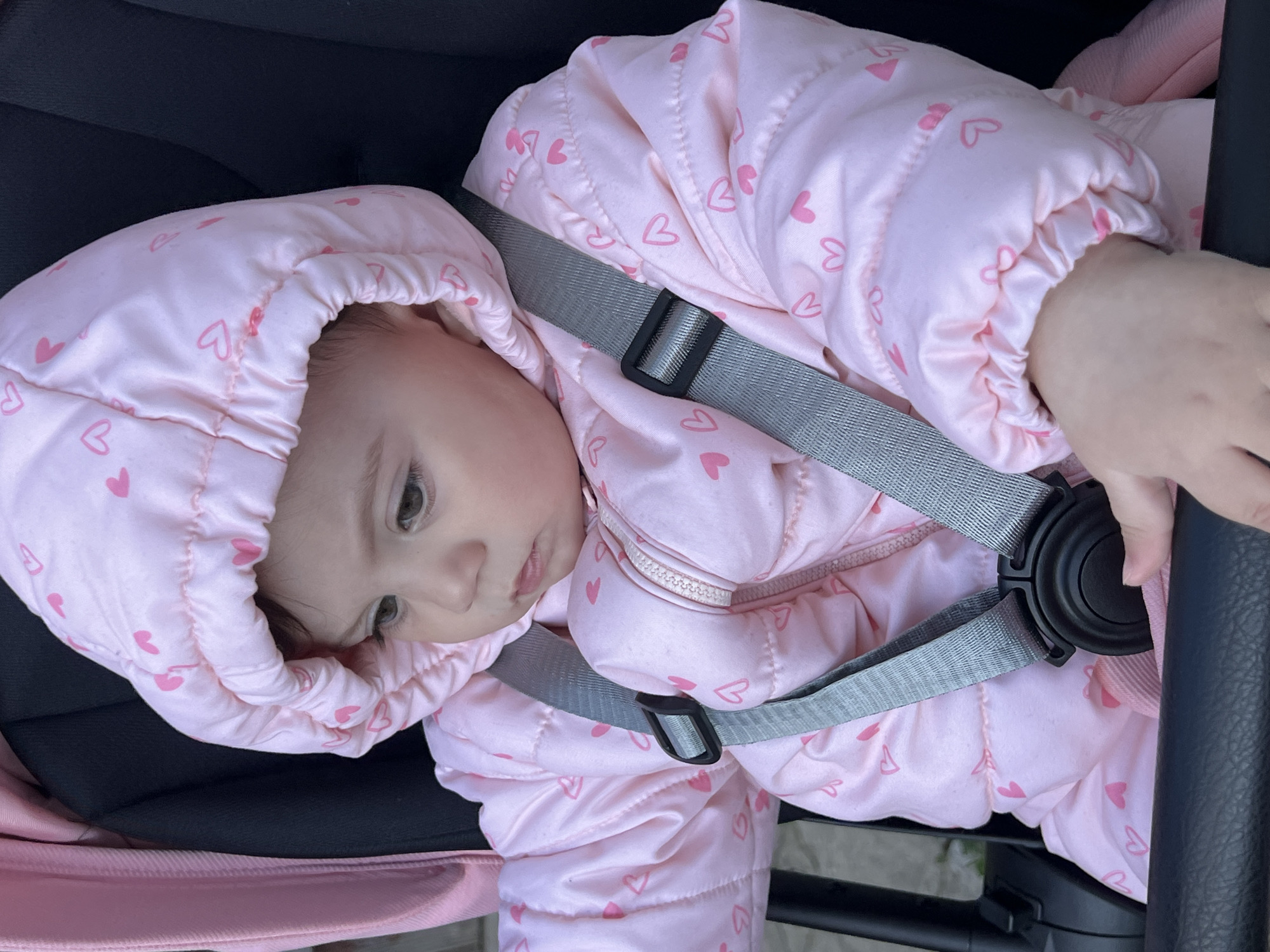 A baby wearing a pink heart-patterned snowsuit is strapped into a stroller. The baby is gazing downwards. The location is not discernable from the image.