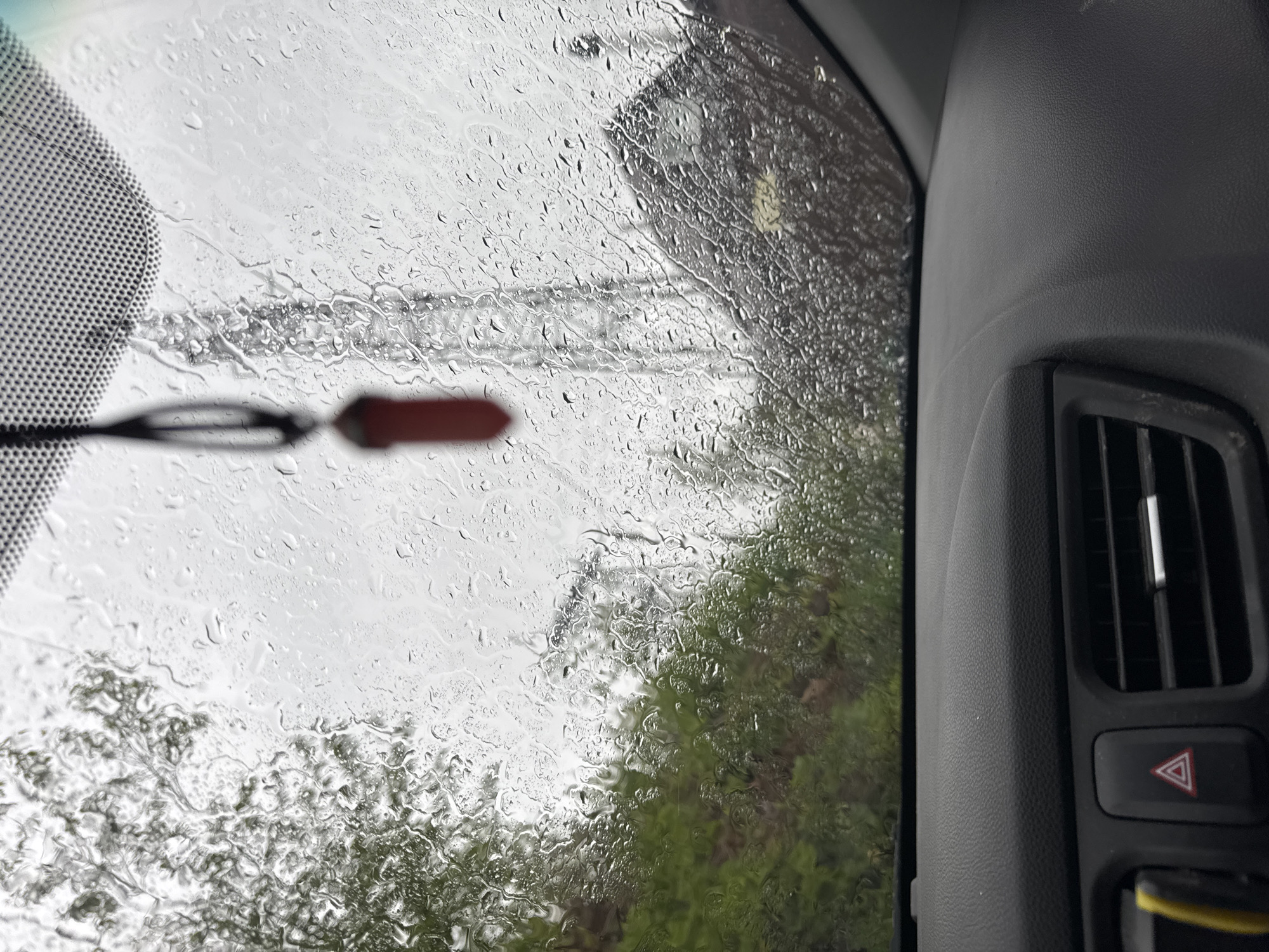 The image shows a view from inside a vehicle on a rainy day, possibly in Chaikovsky, Russia. The foreground features the interior of the vehicle, specifically the dashboard with an air vent and hazard light button. Raindrops obscure the view through the windshield, revealing blurred shapes of a building, power lines, and foliage outside. A blurry object, possibly a rearview mirror component, is visible in the upper left corner. The lighting suggests overcast conditions.