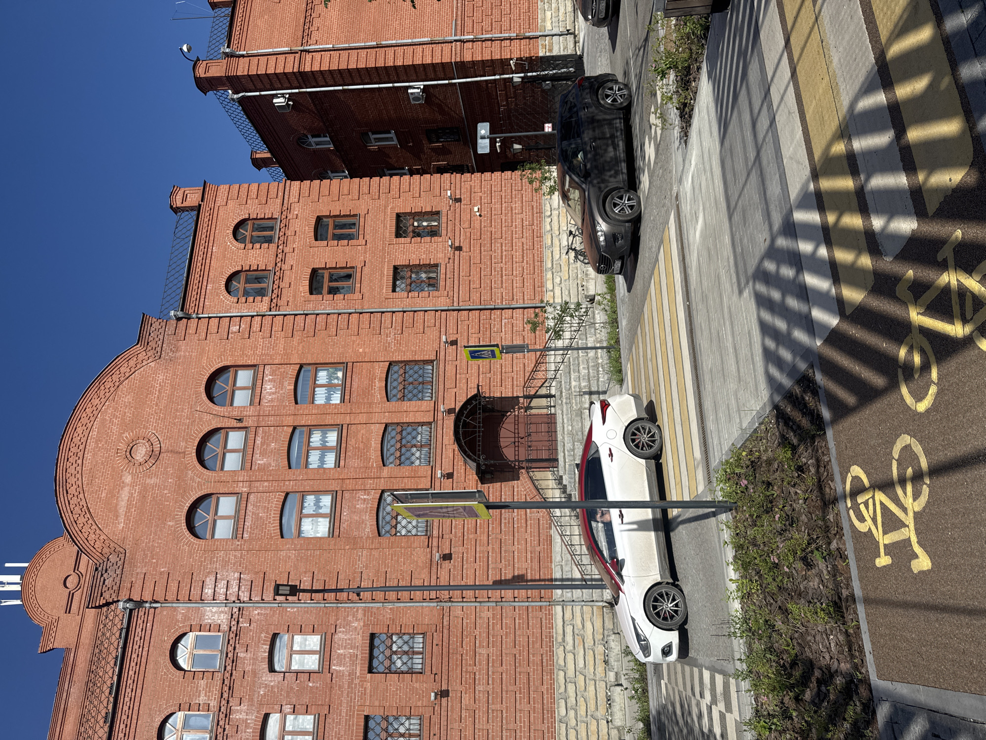 The image shows a street scene in Yekaterinburg, Russia, under a clear blue sky. The dominant feature is a large, multi-story brick building with arched and rectangular windows. To the right of the building, a pedestrian crossing with yellow and white stripes is visible, adjacent to a dedicated bike lane marked with a bicycle symbol.
Two cars are parked on the street. A dark-colored sedan is positioned near the building, and a white sedan is parked closer to the pedestrian crossing. The presence of the bike lane and pedestrian crossing suggests an urban environment designed for mixed-use traffic.
The time of day appears to be daytime, likely late morning or afternoon, given the strong shadows cast by the building and a railing onto the pedestrian area. The weather is sunny and clear, contributing to the bright illumination of the scene. The architecture of the brick building, with its decorative elements and multiple window styles, hints at a historical or repurposed industrial structure. The overall atmosphere is one of a typical urban day, with elements of both historical architecture and modern infrastructure for transportation. No text is clearly legible in the image.