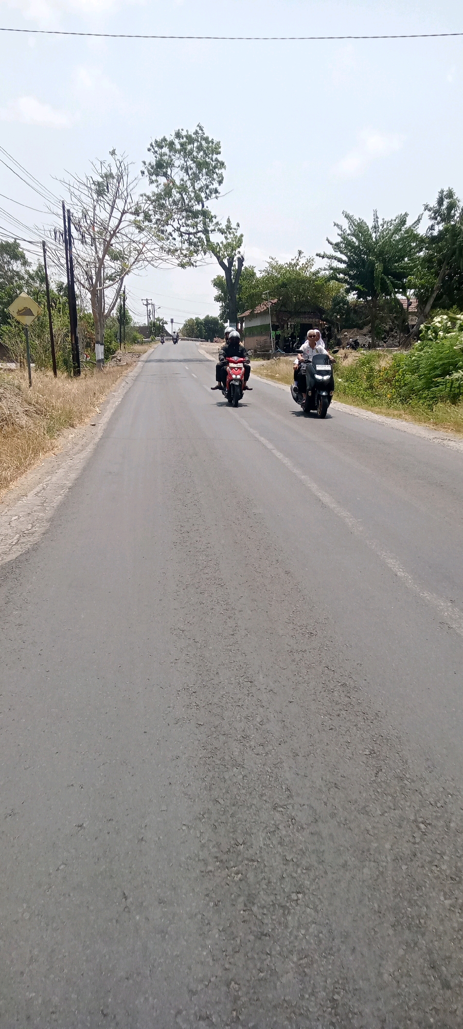 A vertical, eye-level shot captures a rural asphalt road with sparse vegetation and a clear sky. Two motorcyclists are visible. The first, closer to the foreground, is on a red scooter, with two individuals wearing helmets. The second motorcyclist is on a black scooter, also with two individuals wearing helmets, further down the road. To the left, a yellow diamond-shaped road sign with a black incline symbol indicates a steep road. Utility poles and power lines line the left side of the road. Dry, light-brown grass covers the verge. In the background, a few trees with green foliage stand, and a small, traditional Indonesian house with a white and green facade is visible on the right. The road appears to be in the Keteguhan area of Indonesia, a location known for its rural landscapes and agricultural activities.