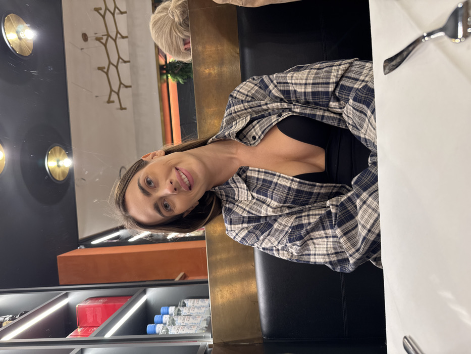 A woman is seated centrally in a restaurant or bar in Verona, Italy, facing the camera with a slight smile. She has long, light brown hair and is wearing an unbuttoned blue and off-white plaid shirt over a deep v-neck black top. She sits at a white table, with a silver fork visible on the right side of the frame. Behind her is a dark upholstered booth and a golden-toned partition. To the left, a shelf unit with illuminated LED strips displays red boxes and multiple clear bottles with blue and white labels. In the background, behind the partition, the top of a blonde person's head with hair tied up is partially visible. The ceiling is dark with circular gold-accented light fixtures and a decorative, branching gold chandelier. An orange wall section and hints of green foliage are also visible further in the background, suggesting interior decoration.