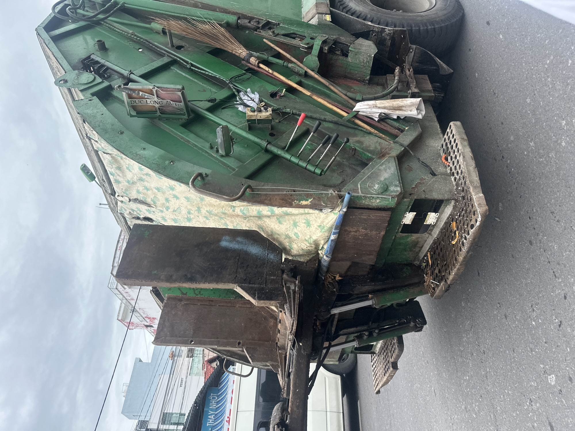 The image depicts a green garbage truck in Bac Nha Trang Ward, Vietnam. The truck is parked on an asphalt road with buildings visible in the background under a cloudy sky. Various tools, including a broom and screwdrivers, are stored on the truck's upper surface. There is a sign that says “DUC LONG AUTO”. A portion of a sign that reads "THAY NHOT" is visible near the truck.