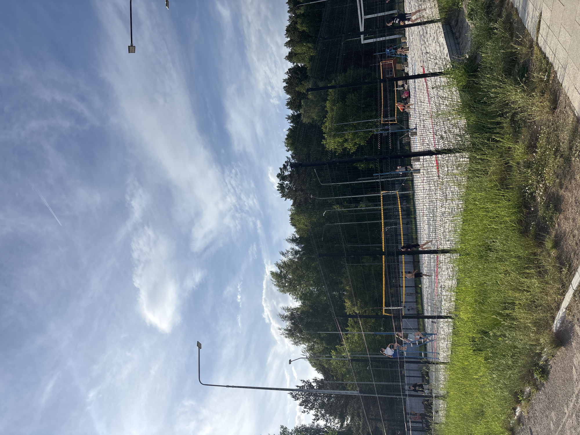 A sideways shot captures a scene featuring a beach volleyball court. The court, lined with black netting and yellow poles, has white sand and red boundary markers. Several figures are positioned on the court, some appearing to be in motion, perhaps playing volleyball. A row of trees borders the court, creating a natural backdrop. Tall streetlights stand alongside the court, extending upwards into a sky filled with wispy clouds. Overgrown grass lines the edge of the court where it meets a paved sidewalk.
