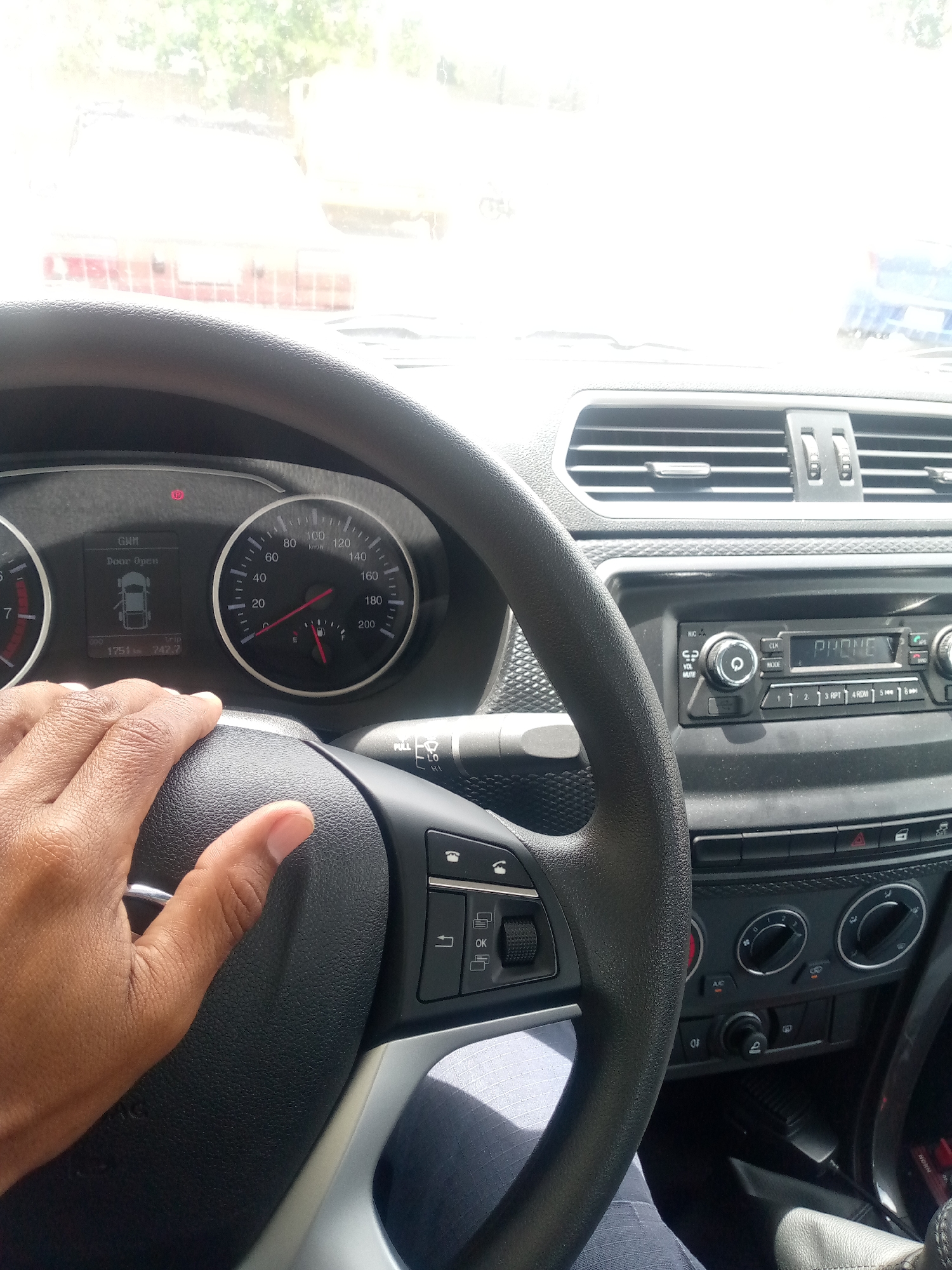 The image shows the interior of a car, specifically the driver's seat area. A hand rests on the black steering wheel, with a clear view of the dashboard and instrument panel. The speedometer reads approximately 10 km/h, and the odometer shows 1751 km. The radio is visible, along with various buttons and controls for the car's functions. The view through the windshield is washed out with light, but some outlines of buildings are discernible, suggesting an outdoor environment.
Based on the text in the image "Door Open" and "1751" combined with the other text, the car is likely a DFSK, and the location is most likely Porlamar, Venezuela.