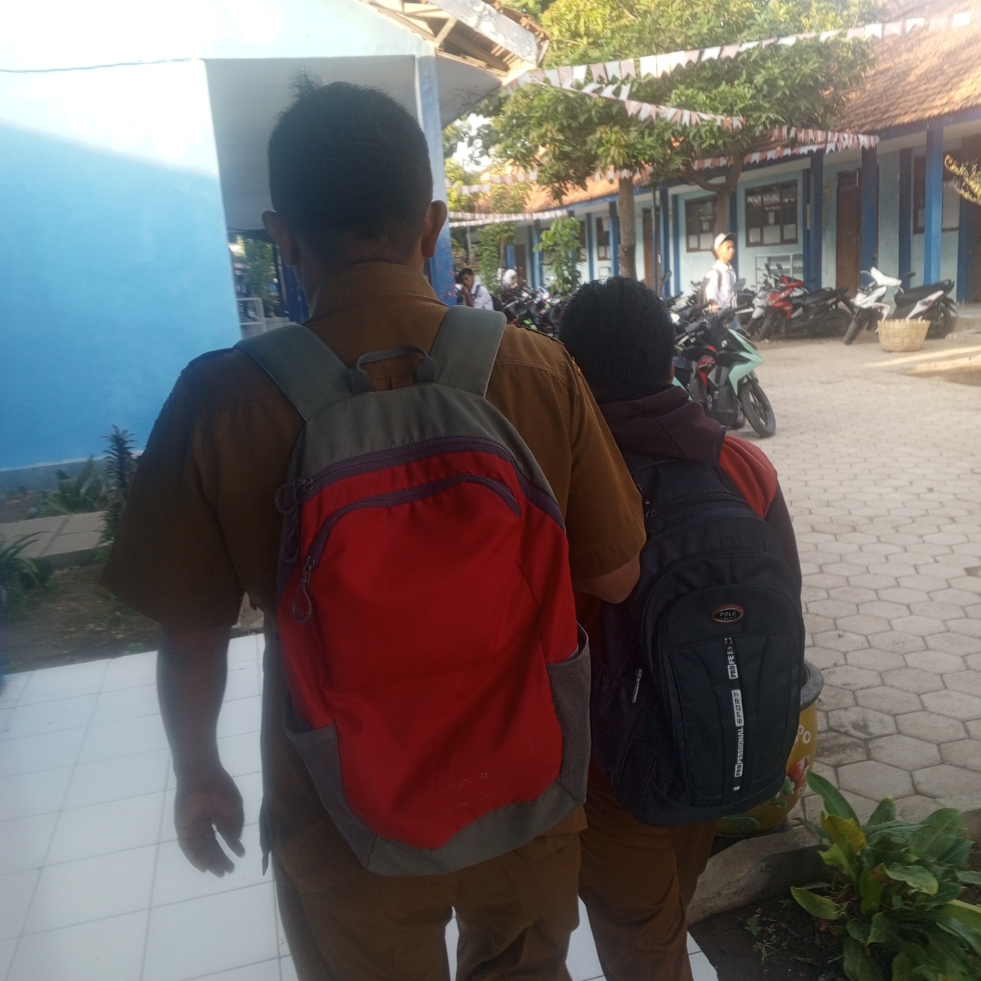 Two individuals, viewed from behind, stand on a tiled pathway adjacent to a building. Both are wearing brown trousers. The person on the left has a tan-colored shirt and is carrying a red backpack with gray accents. The person on the right has a dark hooded garment and is carrying a black backpack. Behind them, a building with blue trim and a tiled roof is visible, along with trees and parked motorcycles. The scene appears to be outdoors in a school or institutional setting.