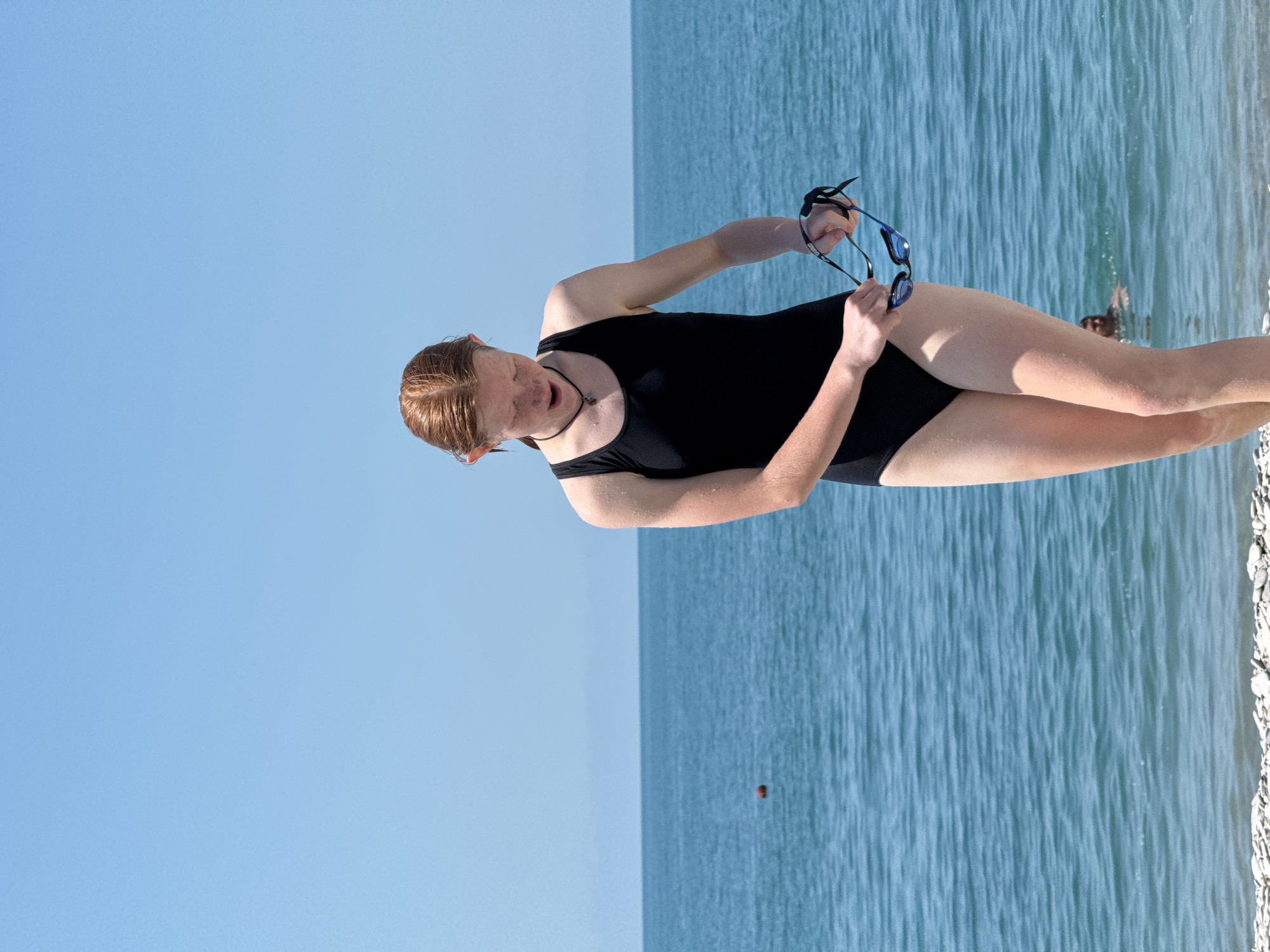 A young Caucasian female with red hair, wearing a black one-piece swimsuit and a black necklace, stands on a pebble beach facing the sea. She is holding a pair of blue and black swimming goggles. Her mouth is slightly open, and her eyes are looking down at the goggles. The sea is calm and blue, with a few ripples. In the background, another person is partially visible in the water. The sky is clear and blue. The image appears to be taken on a sunny day at the beach in Nebug, Russia, a resort town on the Black Sea coast.