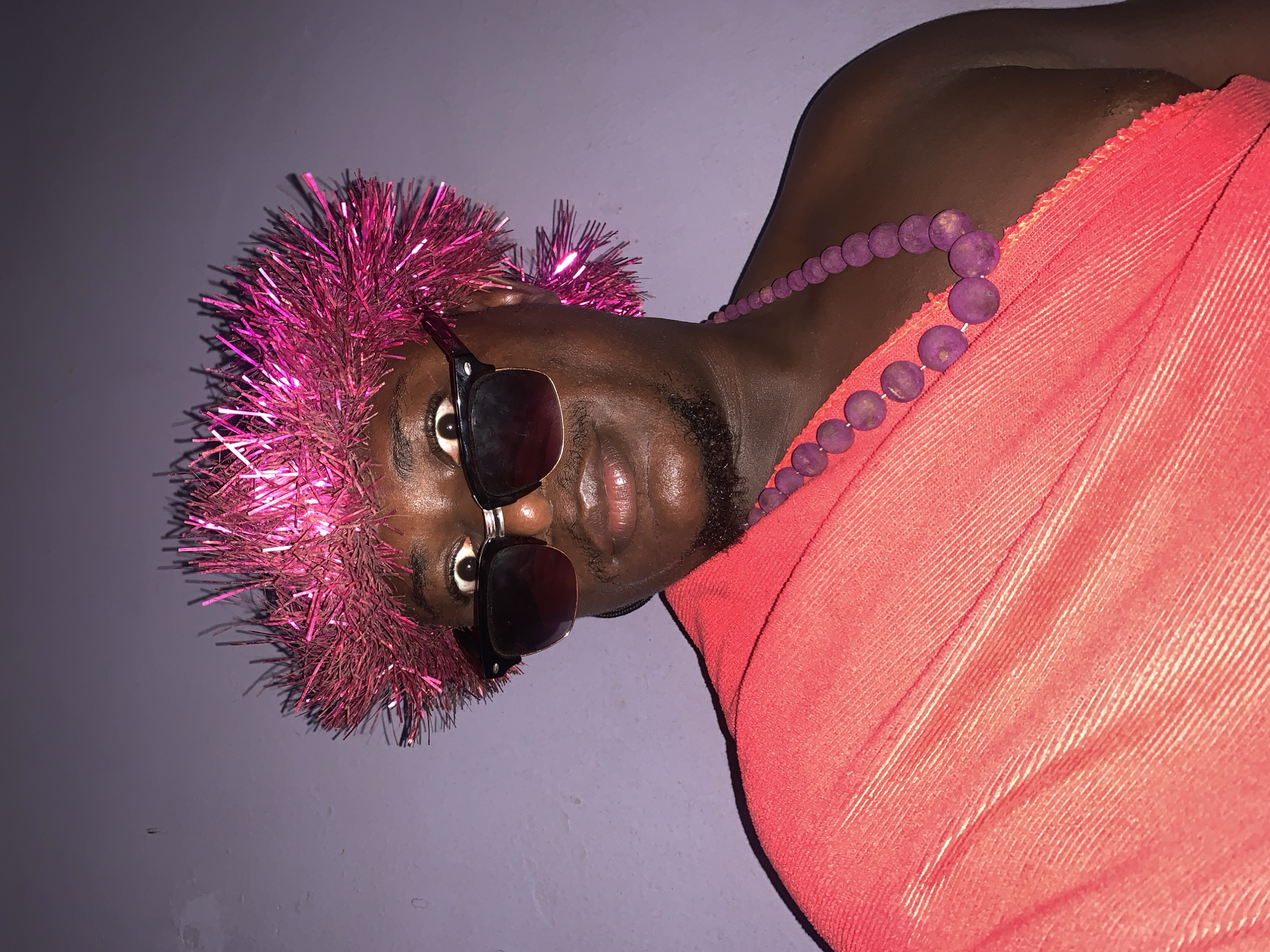 A person is shown from the chest up, looking directly at the camera. They are wearing a pink tinsel headdress and dark sunglasses. A coral-colored garment is draped over their left shoulder, and a purple beaded necklace is visible. The background is a plain, light purple wall. The scene appears to be indoors, likely in a residence. The person's expression is surprised, with wide-open eyes. The lighting is somewhat dim, casting shadows on the wall. The image was taken in Akwanga, Nigeria.