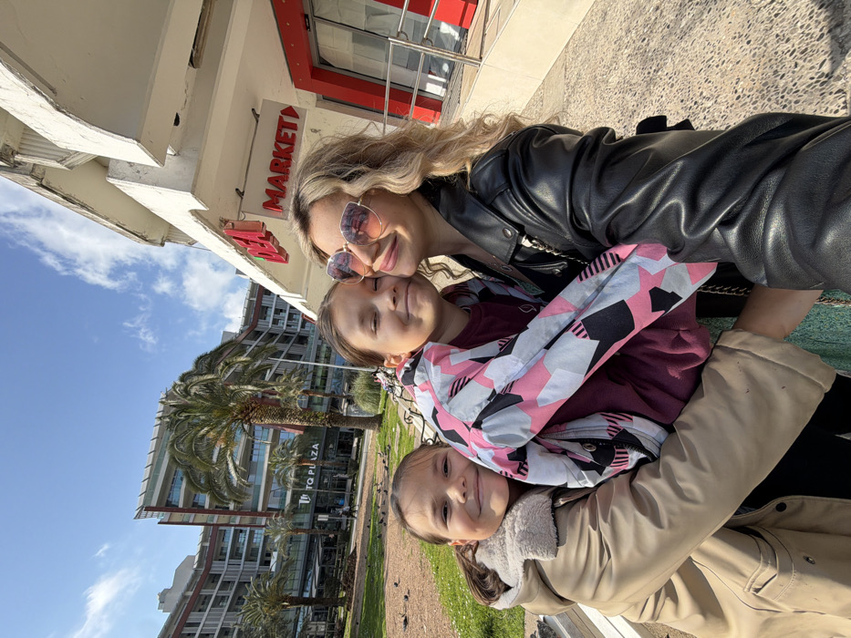 This image depicts three females—an adult and two children—in a close embrace, posing for a selfie outdoors on a sunny day. The adult female on the right, with blonde, wavy hair, wears a black leather jacket and large, reflective pink-tinted sunglasses, smiling towards the camera. Her left arm is around the central child. The central child, wearing a multi-colored geometric-patterned jacket over a maroon top, smiles with closed eyes, leaning into the adult. The child on the left, wearing a tan jacket with a lighter-colored collar, also smiles with closed eyes, embracing the central child.
The background shows an urban environment. To the left and center, a grassy park area with palm trees and scattered pigeons is visible. Behind the park stands a prominent multi-story building identified by "TQ PLAZA" signage. To the right, a white building features red neon signs reading "DEA" and "MARKET" above a red-framed entrance. The sky is clear blue with scattered white clouds, indicating bright daylight.
This scene is located in Budva, Montenegro, specifically in the vicinity of the TQ Plaza complex and a DEA Market, which are recognizable landmarks in the city.