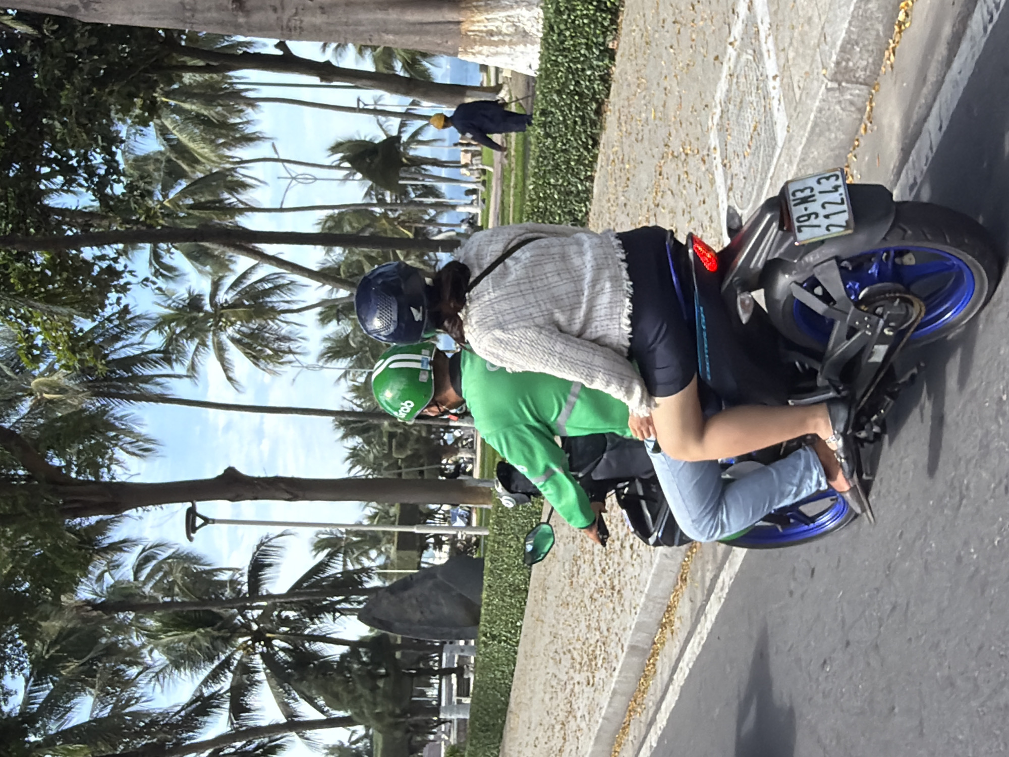 A Grab motorcycle driver, wearing a green jacket and helmet, is operating a motorcycle on a street in Nha Trang Ward, Vietnam. A female passenger, wearing a light-colored patterned jacket and dark pants, is seated behind him. They are riding on a paved road, with a sidewalk and landscaping, including palm trees and bushes, to their right. In the background, several palm trees line the street, and a large, dark rock formation is visible. Further in the distance, the ocean can be seen, with a person in a blue jacket and yellow helmet walking near the shore. The motorcycle has a license plate that reads "79-N3 212.43". The sky is partly cloudy.