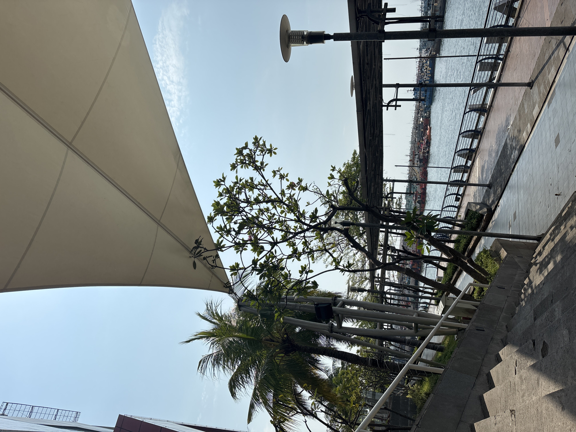 The image shows an outdoor scene in North Jakarta, Indonesia, oriented vertically. The sky is blue with wisps of clouds. A large, white fabric shade structure dominates the upper left portion. Trees, including a palm tree, are present below the shade. A row of thin, silver metal poles lines the walkway near the water. The water is visible in the background with docked ships. A grey stone staircase occupies the lower right corner.