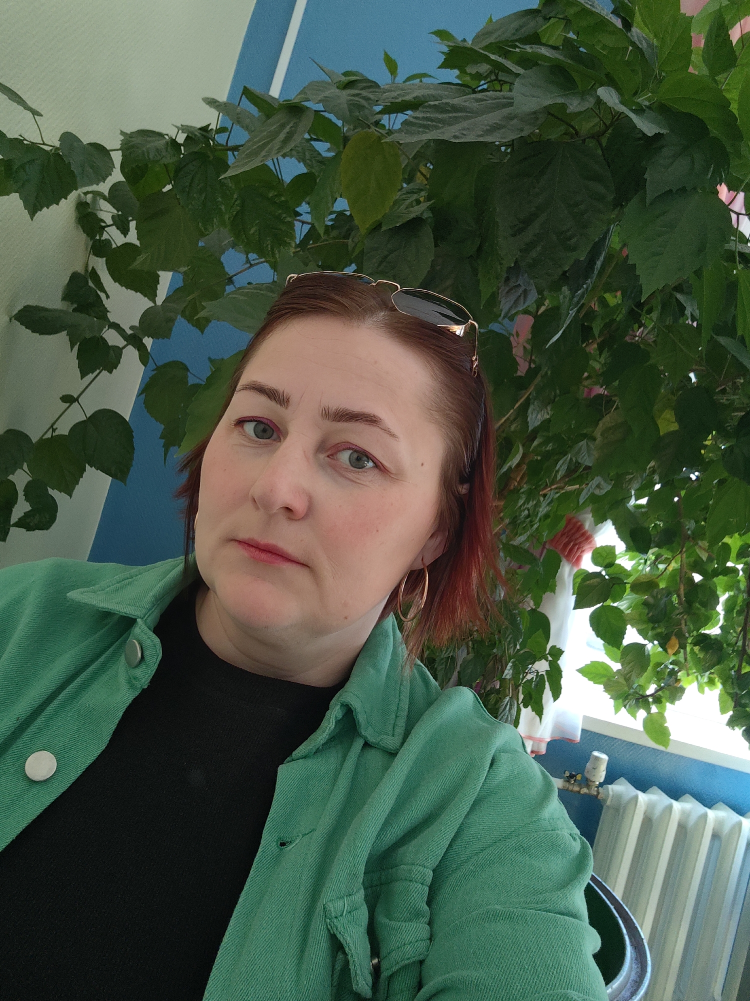 A middle-aged woman with reddish-brown hair and fair skin is the focus of a selfie-style photograph. She wears a green jacket over a black top and gold hoop earrings, and her sunglasses are perched on top of her head. The woman has a serious expression and looks directly at the camera. Behind her is a lush green leafy plant, and a blue and white striped wall is partially visible in the background. Further in the background, a radiator is visible. The photograph appears to have been taken indoors, possibly in a home or office in Chaikovsky, Russia.