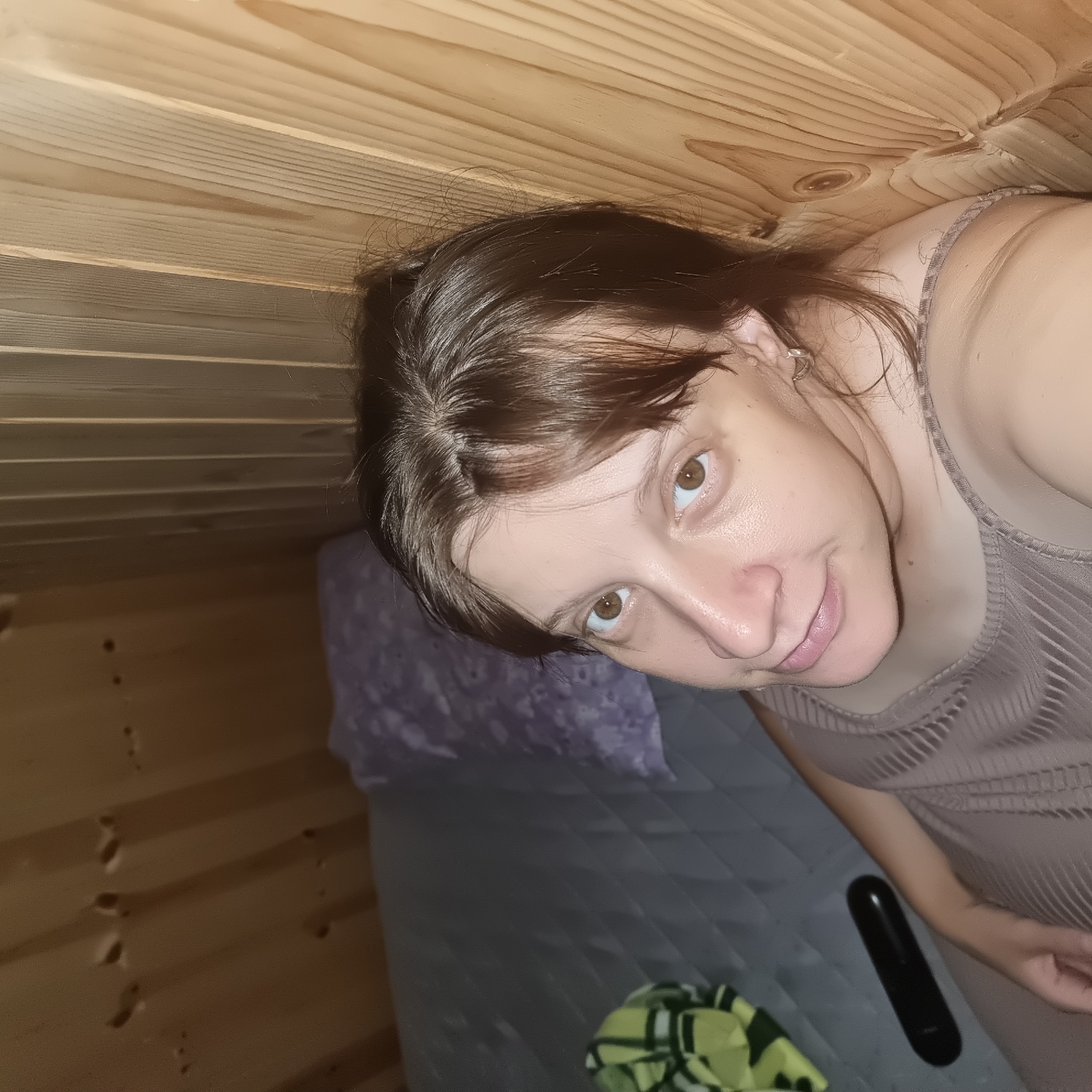 A woman with brown hair and brown eyes is looking up at the camera. She is wearing a sleeveless brown dress. She is sitting on a mattress that is next to a wooden wall. There is a pillow on the mattress and a green and yellow blanket. A black remote control is on the mattress. The scene is indoors, in what appears to be a small room with wooden paneling on the walls and ceiling.