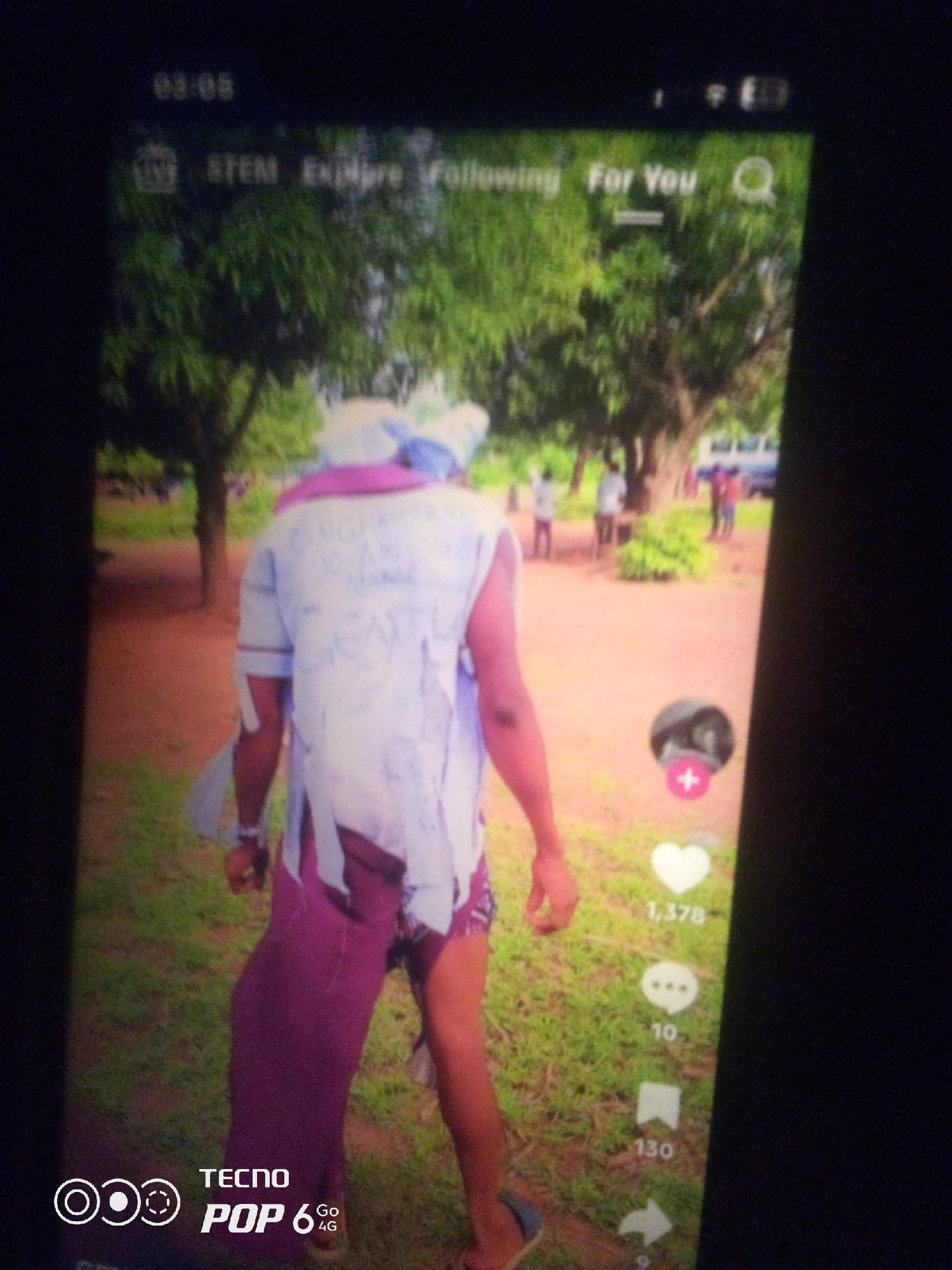 The image captures a full-body view of a person standing in an outdoor environment with a slightly blurry, low-resolution effect, suggesting it was taken with a mobile phone. The person is facing away from the camera, wearing a torn, light-colored shirt with purple accents and a matching purple skirt or garment wrapped around their legs. Text is visible on the back of the shirt, but it is largely illegible due to the image quality. The background features trees and a dirt path, with several other individuals visible in the distance, some standing near a white vehicle. The foreground shows a patch of grass. A mobile phone interface is superimposed on the image, displaying icons for likes, comments, and shares, with numerical counts associated with them. The top of the screen shows the time "01:05" and navigation elements like "STEM," "Explore," "Following," and "For You." At the bottom left corner, a "TECNO POP 6 4G" logo is present. The specific location in the city of Howayi, Nigeria cannot be determined from the visual information provided in the image.