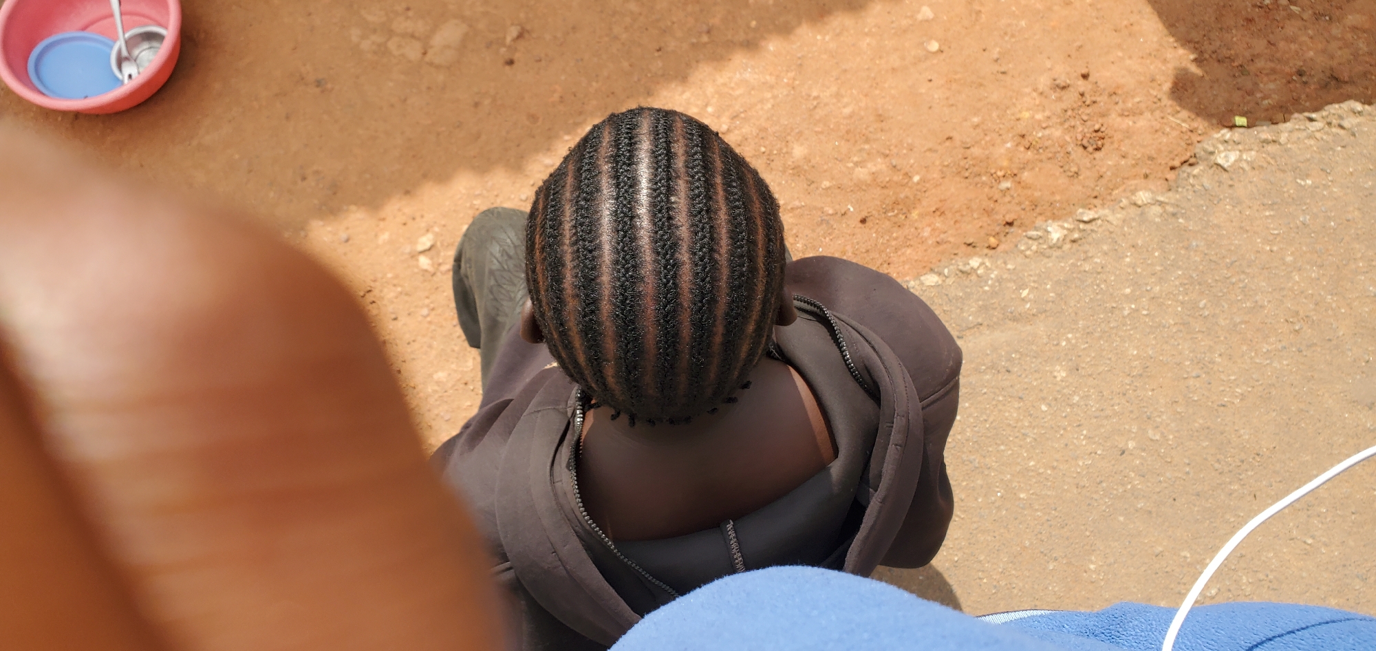 A high-angle shot captures a close-up view of a person's head from the crown, showcasing a meticulously braided hairstyle. The braids are arranged in uniform horizontal lines across the scalp, creating a striped effect with alternating dark and lighter brown hues. The individual is wearing a dark-colored zippered jacket, with the zipper partially open. A portion of a light blue garment is visible on the left side of the frame, extending from the bottom edge. The background consists of dry, sandy ground, suggesting an outdoor setting. To the upper right, a pink plastic bowl with a blue lid and a spoon inside is partially visible. The lighting indicates it is daytime, with shadows cast on the ground. The image is taken from an elevated perspective, with a blurred foreground element that appears to be a person's hand or finger. The context of Gura Top, Nigeria, is provided, though no specific landmarks or characteristics within the image directly indicate this location.