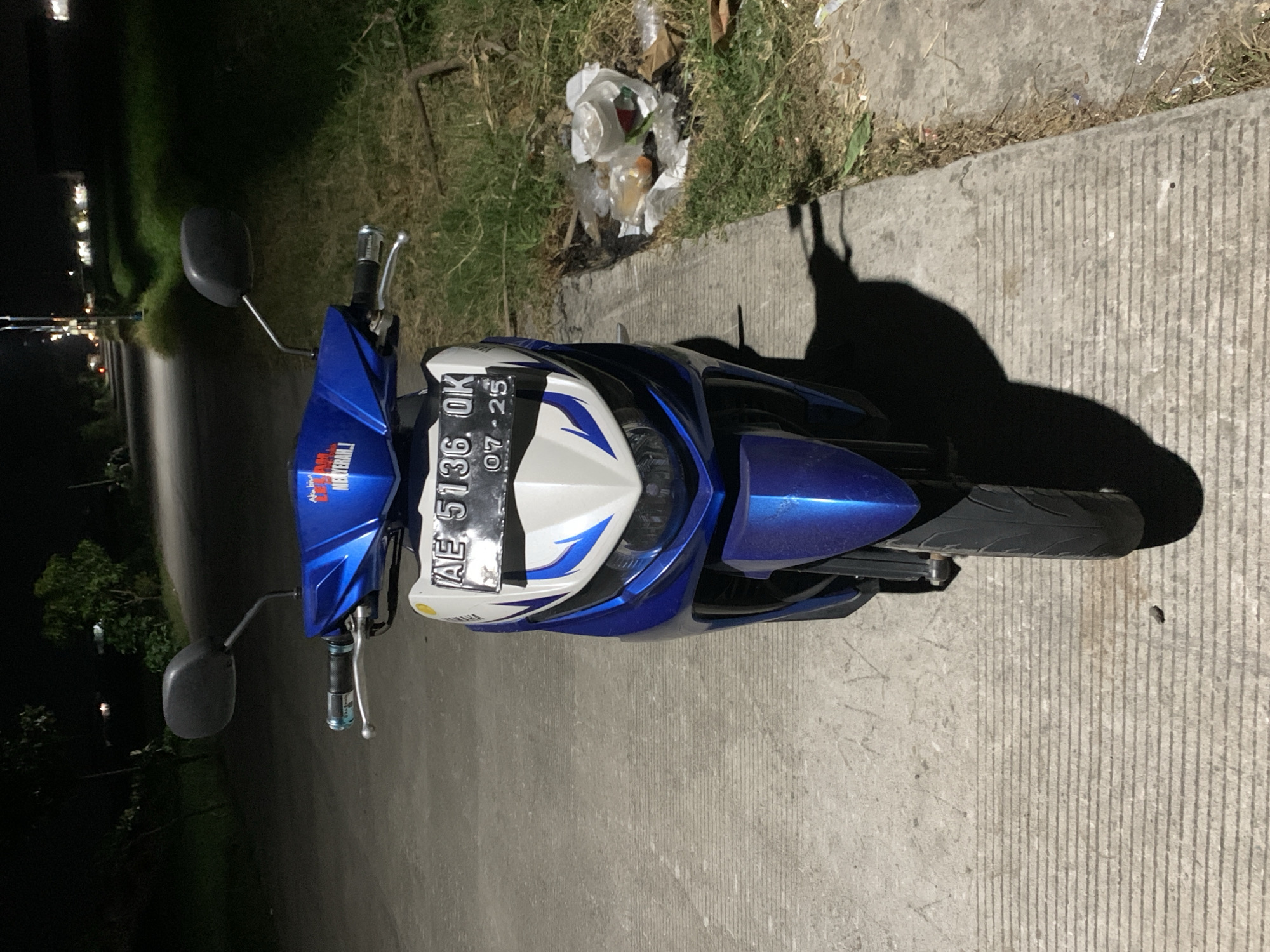 The image depicts a blue and white Yamaha Mio series scooter, parked head-on on a concrete paved surface during nighttime in Jungke, Indonesia. The scooter's front fairing is white with blue and dark blue graphic decals, featuring a "YAMAHA" logo. The handlebar cowl is blue, bearing a red sticker with partially visible Indonesian text, including "MENYERAH!" (surrender). The scooter is equipped with a headlight, two rearview mirrors, and handlebar grips, one with a light blue accent. The front license plate, centrally positioned, reads "AE 5136 OK" with "07-25" (indicating month and year) printed below.
The scooter is situated on a light grey concrete surface characterized by distinct horizontal grooves. To the right of the scooter, a narrow strip of unkempt grass contains discarded refuse, including various plastic bags and what appear to be food containers. A dark, possibly metallic, object is partially embedded in the grass further back. To the left, the concrete surface extends without immediate features.
The background reveals an asphalt road leading into the distance under artificial illumination from multiple streetlights, indicating a nighttime setting. Dark foliage and trees are visible on both sides of the road in the mid-ground and background. Distant lights are discernible, suggesting a settlement or infrastructure beyond the immediate vicinity. No individuals are present in the scene.