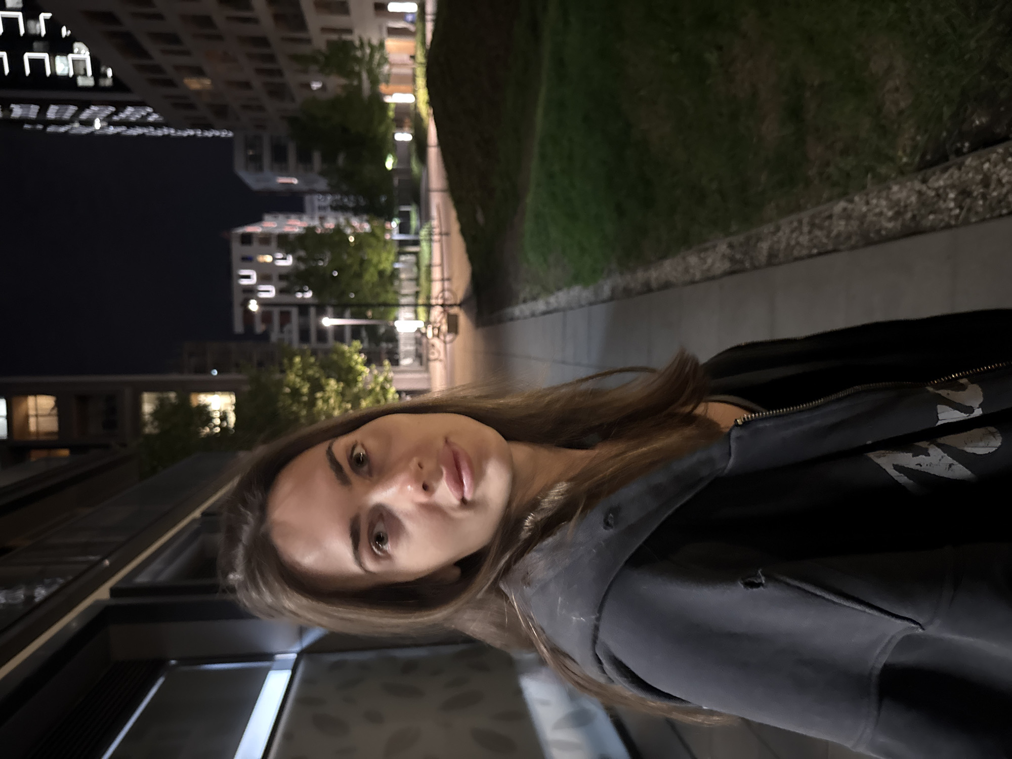 A nighttime photograph shows a woman with long, light brown hair wearing a dark jacket standing on a sidewalk in a city. To her right is a grassy area bordered by a curb. Buildings with illuminated windows are visible in the background. The lighting suggests it is night.