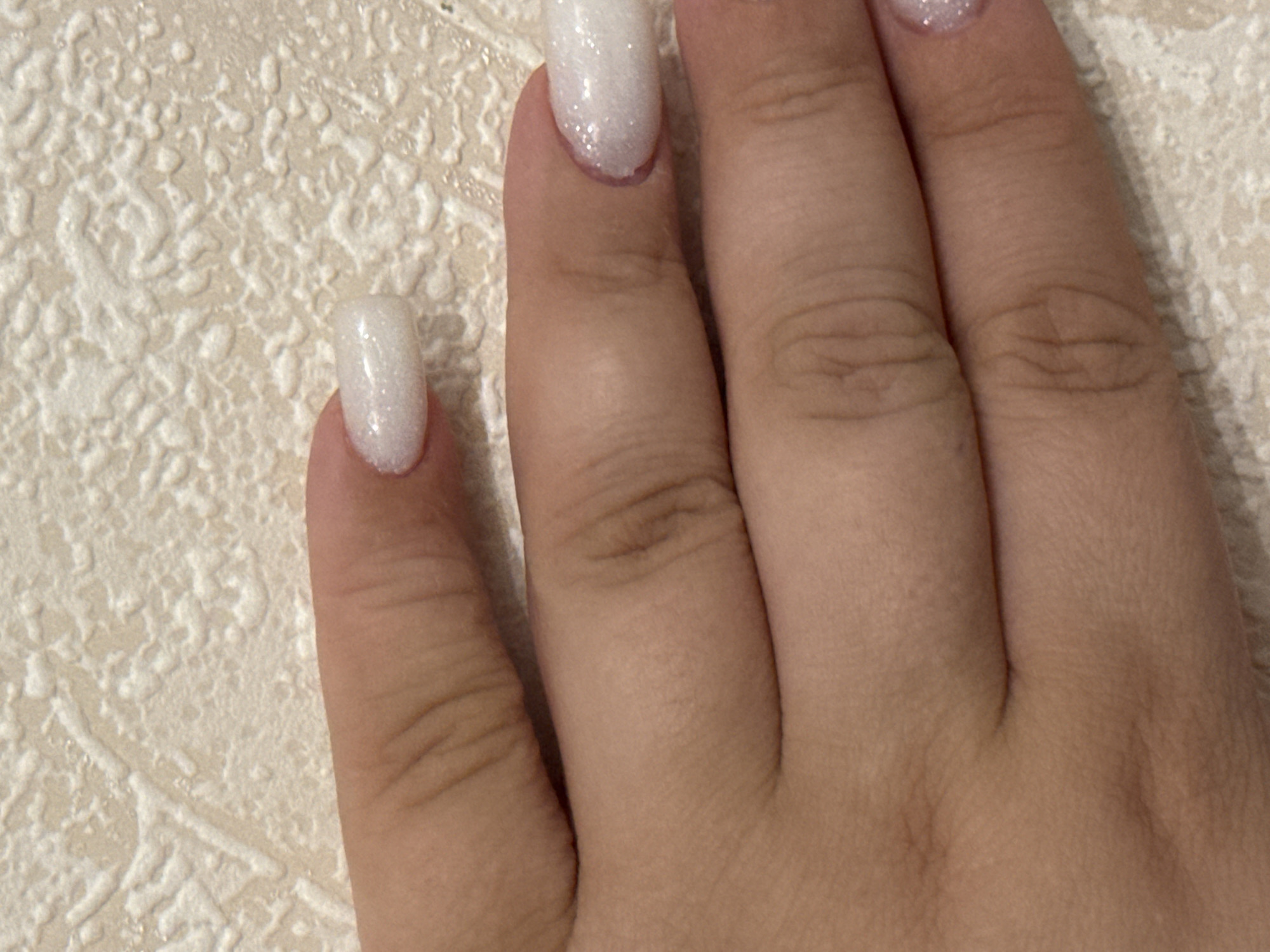 The image displays a close-up view of a person's hand, specifically the fingers and nails, against a textured, off-white background. The fingernails are long, oval-shaped, and appear to be painted with a glittery white polish. The background has a raised, uneven pattern, resembling wallpaper or a textured surface. There are no discernible elements that would indicate a specific location within the city of Saratov, Russia. The image is a static shot, capturing a portion of the hand without any action or interaction.