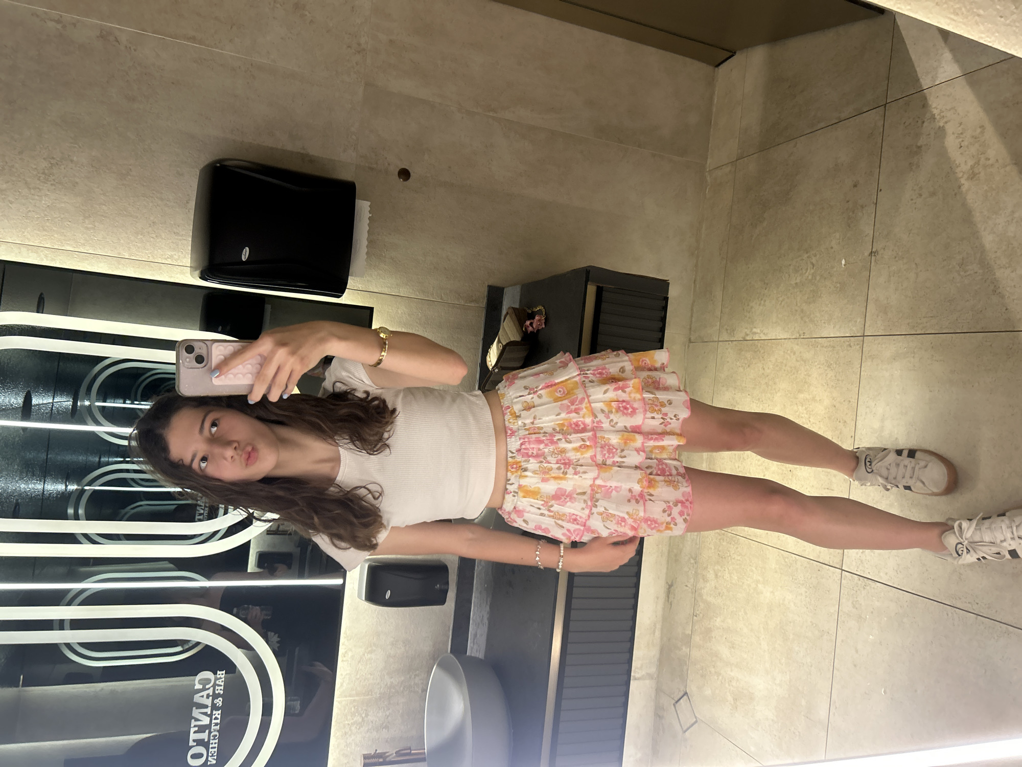 A young woman with long, wavy brown hair is taking a mirror selfie in a modern restroom. She is wearing a white ribbed crop top and a white mini skirt with a pink and yellow floral pattern. She has on white sneakers with black stripes and is accessorized with a gold bracelet and a silver bracelet. She is holding a light pink phone and is making a kissy face. The restroom features a large mirror with backlit vertical stripes, a gray sink with a black countertop, a black paper towel dispenser, and beige tiled walls and floors. The location is not specified as Ruse, Bulgaria.