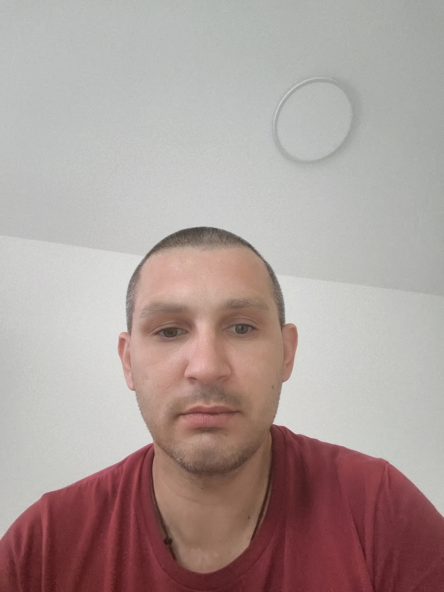 The image shows a close-up, head-and-shoulders shot of a man. He has a shaved head, light brown eyes, and a light stubble. He is wearing a dark red t-shirt. The background is a plain white wall and ceiling, with a circular light fixture visible on the ceiling. There is no indication of the specific location within the city of Wolgast, Germany.