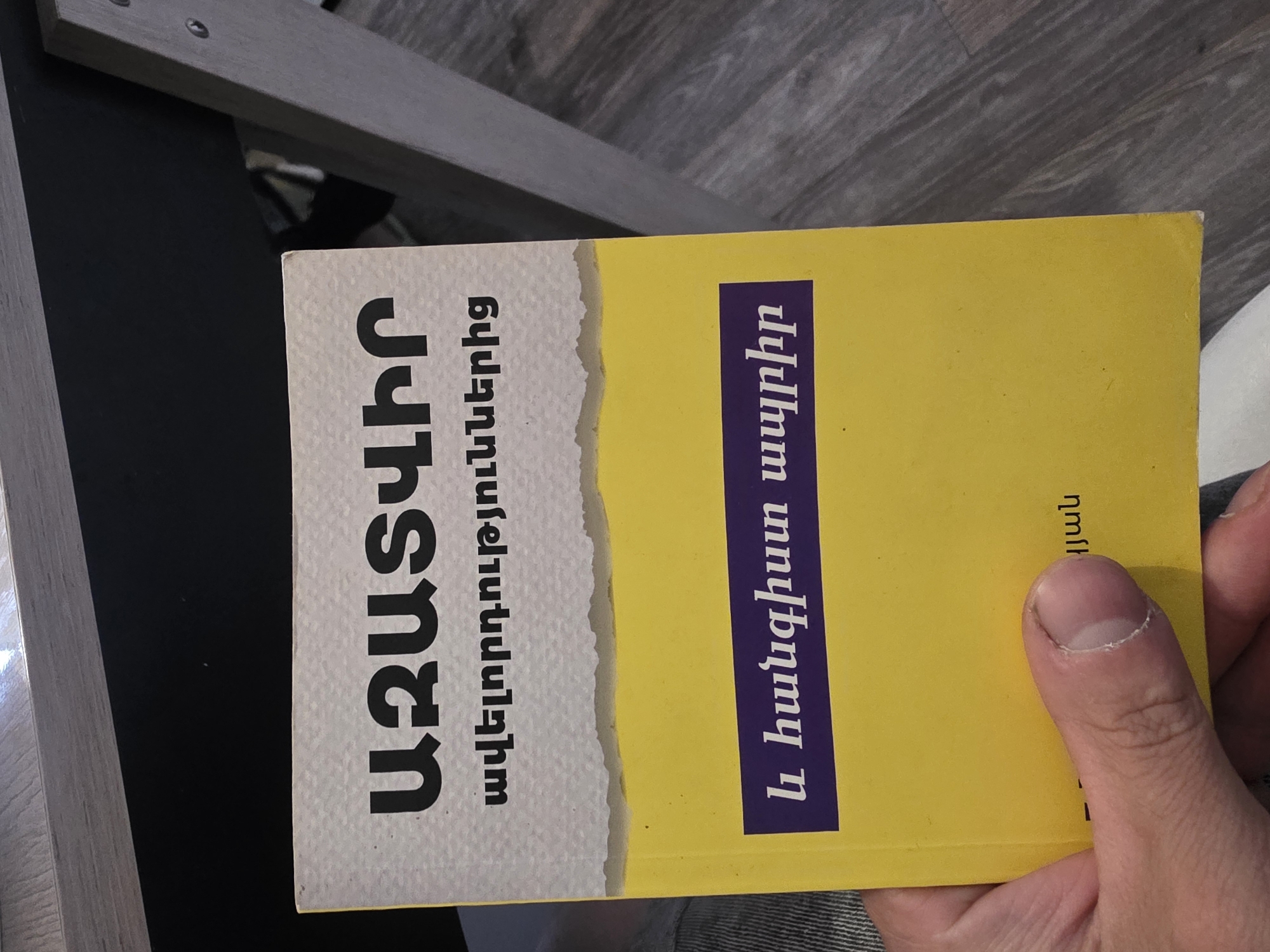 A book with an Armenian title is held by a person's hand. The book has a yellow cover with text in Armenian. The top portion of the book has a white textured background with black text, followed by a jagged line that separates it from the yellow portion. At the bottom of the yellow cover, there is a purple rectangular box with white Armenian text. The book is angled and lies on a surface that appears to be a wooden floor. The lighting suggests it is indoors. There are no visible clues about the specific city or location.
