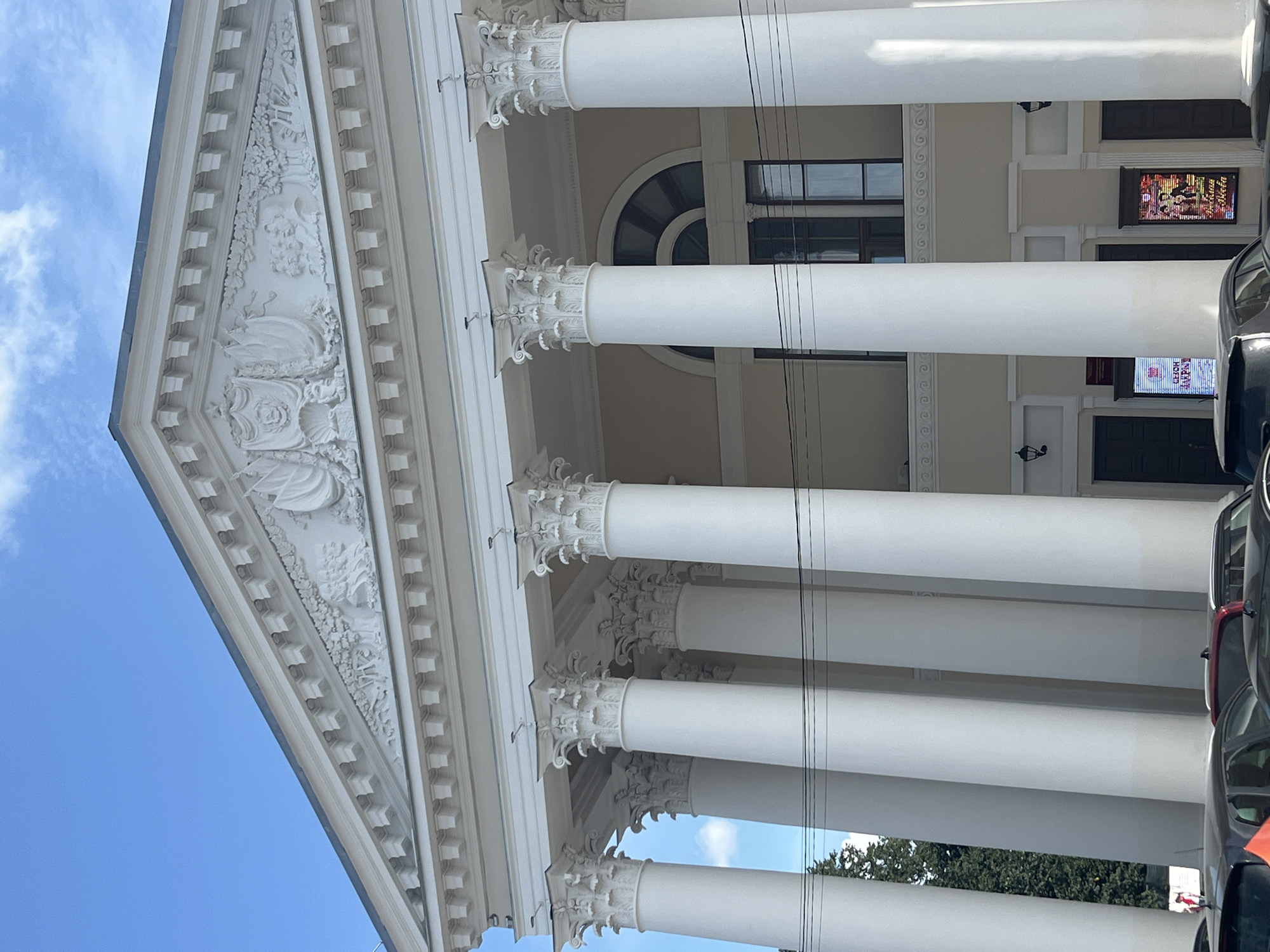 The image shows the exterior of a grand building in Tver, Russia, likely a cultural institution, featuring neoclassical architecture. A prominent facade with a triangular pediment adorned with relief sculptures is visible at the top. Below the pediment, a colonnade of tall, white Corinthian columns supports an entablature. The columns are evenly spaced and create a sense of grandeur and symmetry. Behind the columns, windows and doorways are partially visible, along with some decorative architectural elements. A bright blue sky with scattered clouds forms the background. In the foreground, the tops of several cars are visible, indicating the presence of a street or parking area in front of the building. The lighting suggests it is daytime. The overall impression is of a historic and well-maintained building.