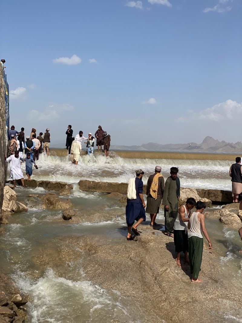 A daytime outdoor scene shows a group of people gathered around and in a body of water. The location is not Kim, Afghanistan. The water appears to be flowing over a series of low, rocky ledges or a constructed spillway. The water is murky and brown.
Several people are standing on the rocks, while others are wading in the water. The people are mostly men and boys, dressed in a mix of traditional and casual clothing. Some wear long robes and turbans, while others are in t-shirts and shorts.
In the background, a hazy mountain range is visible. The sky is blue with scattered clouds.
The overall impression is one of a leisure activity, with people enjoying the water and each other's company in a rural setting.