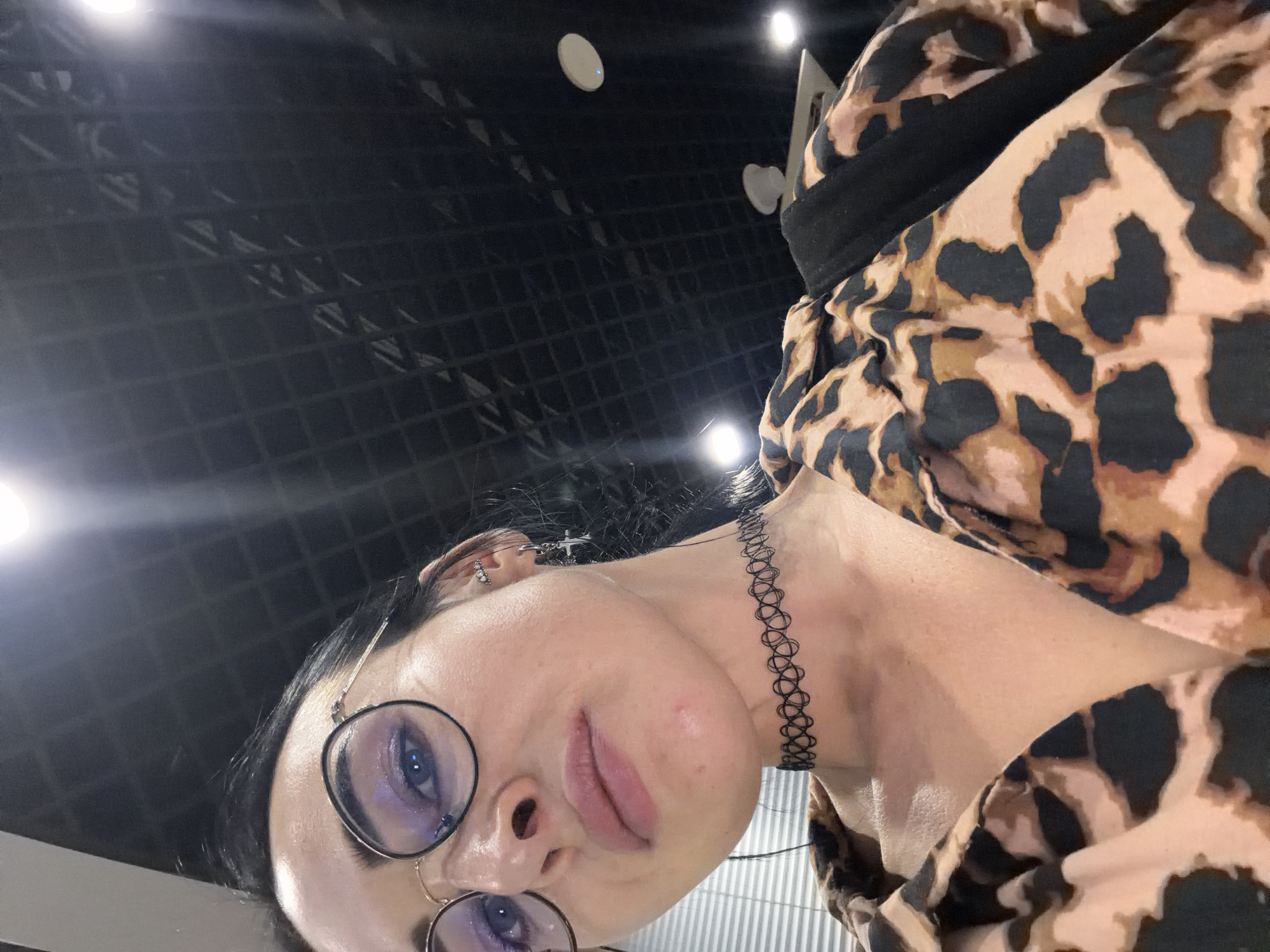 A close-up, low-angle selfie captures a woman in an indoor setting within Saint Petersburg, Russia. The woman, with dark hair and fair skin, directly faces the camera. She wears round, dark-rimmed glasses that partially obscure her light-colored eyes, which exhibit a subtle purple eyeshadow. Her full lips are slightly parted. A black woven choker is visible around her neck. Her left ear (viewer's right) features multiple piercings, including a small silver-colored cross earring and an upper helix piercing with multiple small stones. She is dressed in a leopard-print (tan and black) collared blouse, with a dark strap (possibly from a bag) visible on her upper left shoulder. The background features a dark, grid-patterned ceiling with several bright, circular embedded light fixtures and associated lens flares. A horizontal slatted structure is partially visible on the far left, and some white ceiling-mounted objects are seen on the right.