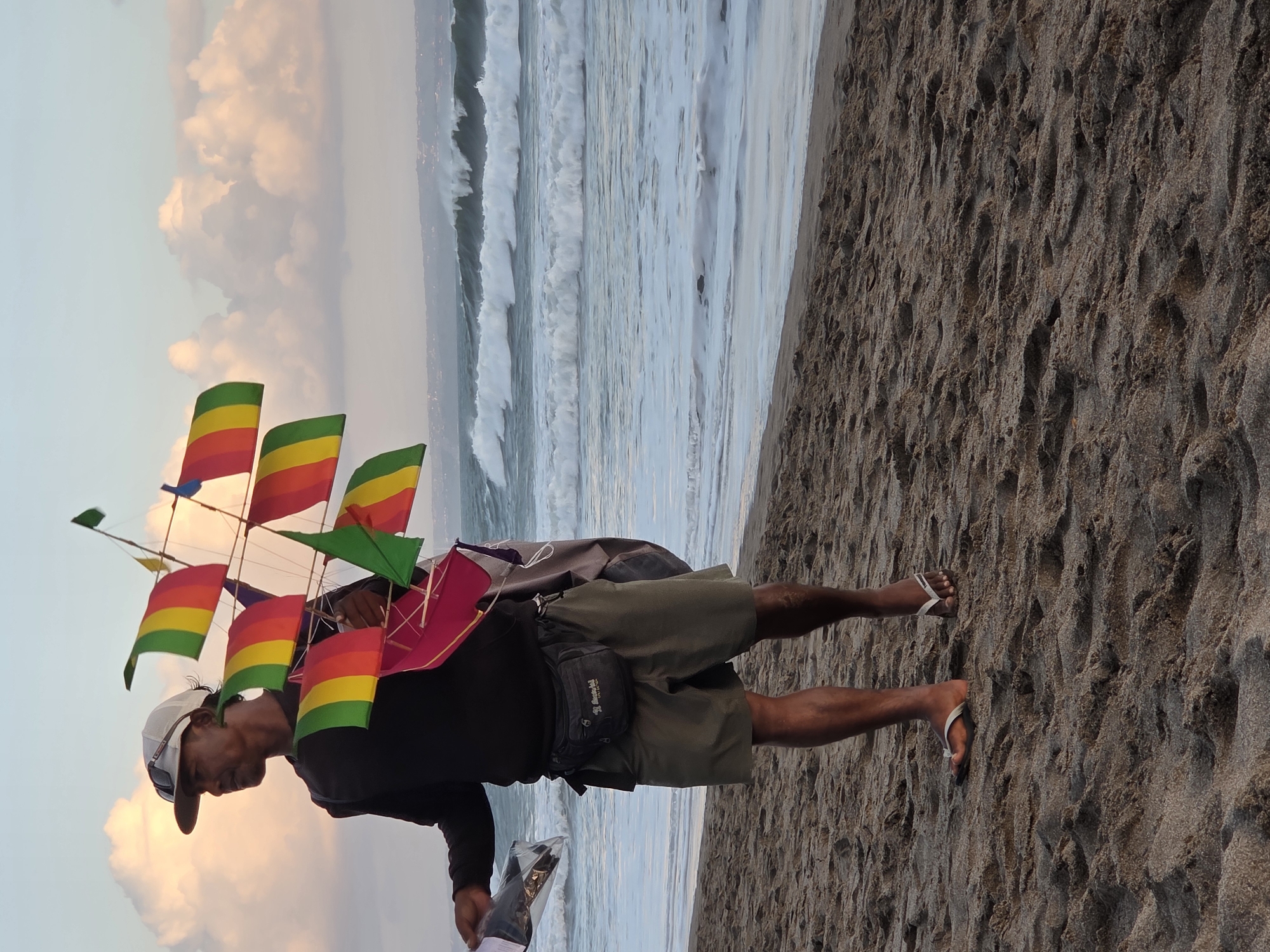 A man stands on a sandy beach with the ocean in the background. He is wearing a black long-sleeved shirt, khaki shorts, a baseball cap, and flip-flops. He is holding a colorful kite shaped like a ship. The kite has red, yellow, and green flags. He also has a small bag hanging from his waist. The waves are crashing on the shore, and the sky is filled with clouds. This scene is likely in North Kuta, Indonesia.
