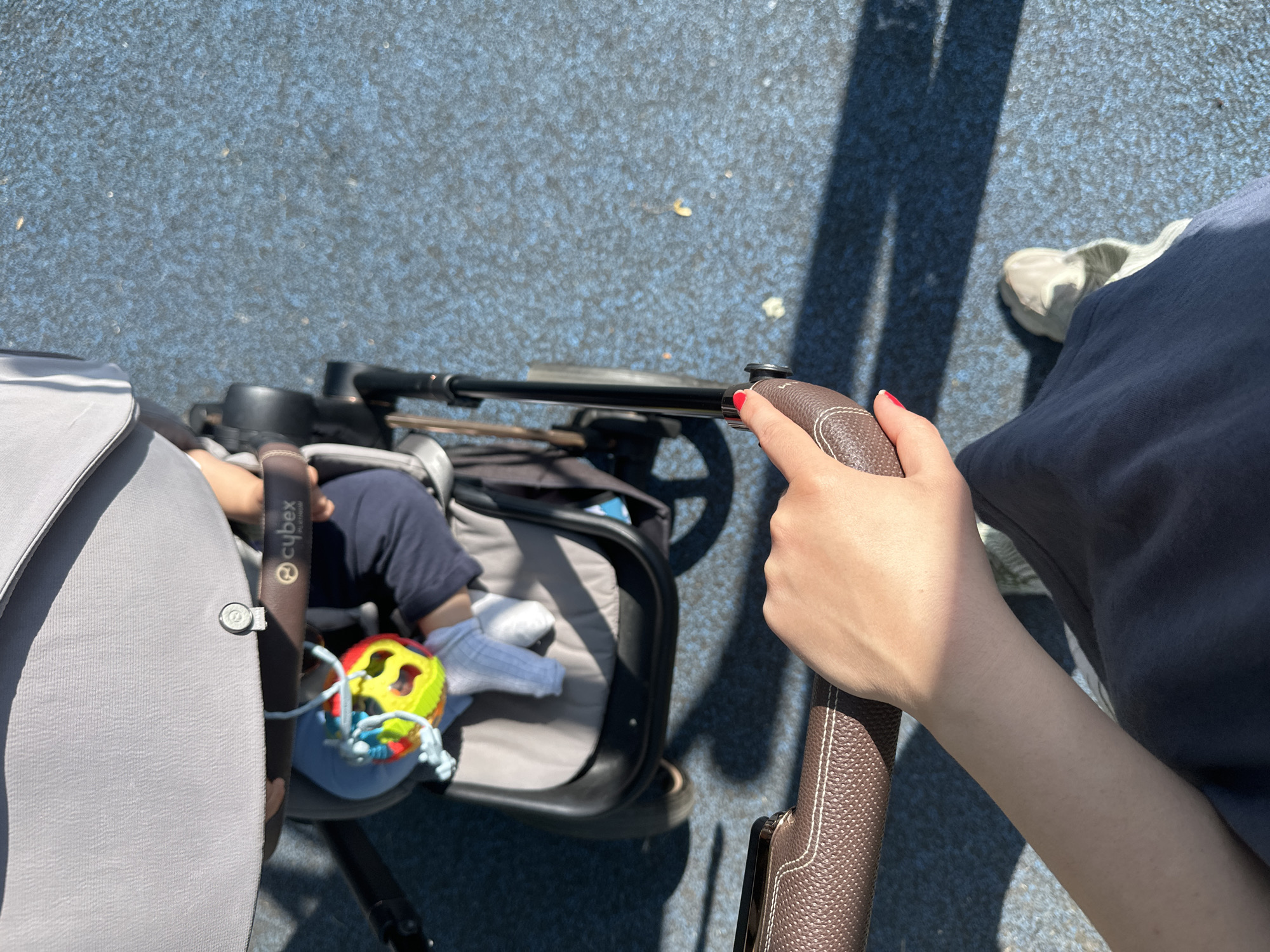 An overhead shot shows a person's hand and forearm gripping the leather-covered handlebar of a stroller. The stroller contains a baby, who is wearing a navy blue outfit and white socks, and is reaching for a colorful rattle toy. The stroller itself is gray and black, with a Cybex logo visible on the handlebar. The stroller is on a blue rubberized surface, likely a playground or park area. The sunlight casts shadows on the blue surface. There is no discernible information within the image to identify the location as the city of Leeds, United Kingdom.